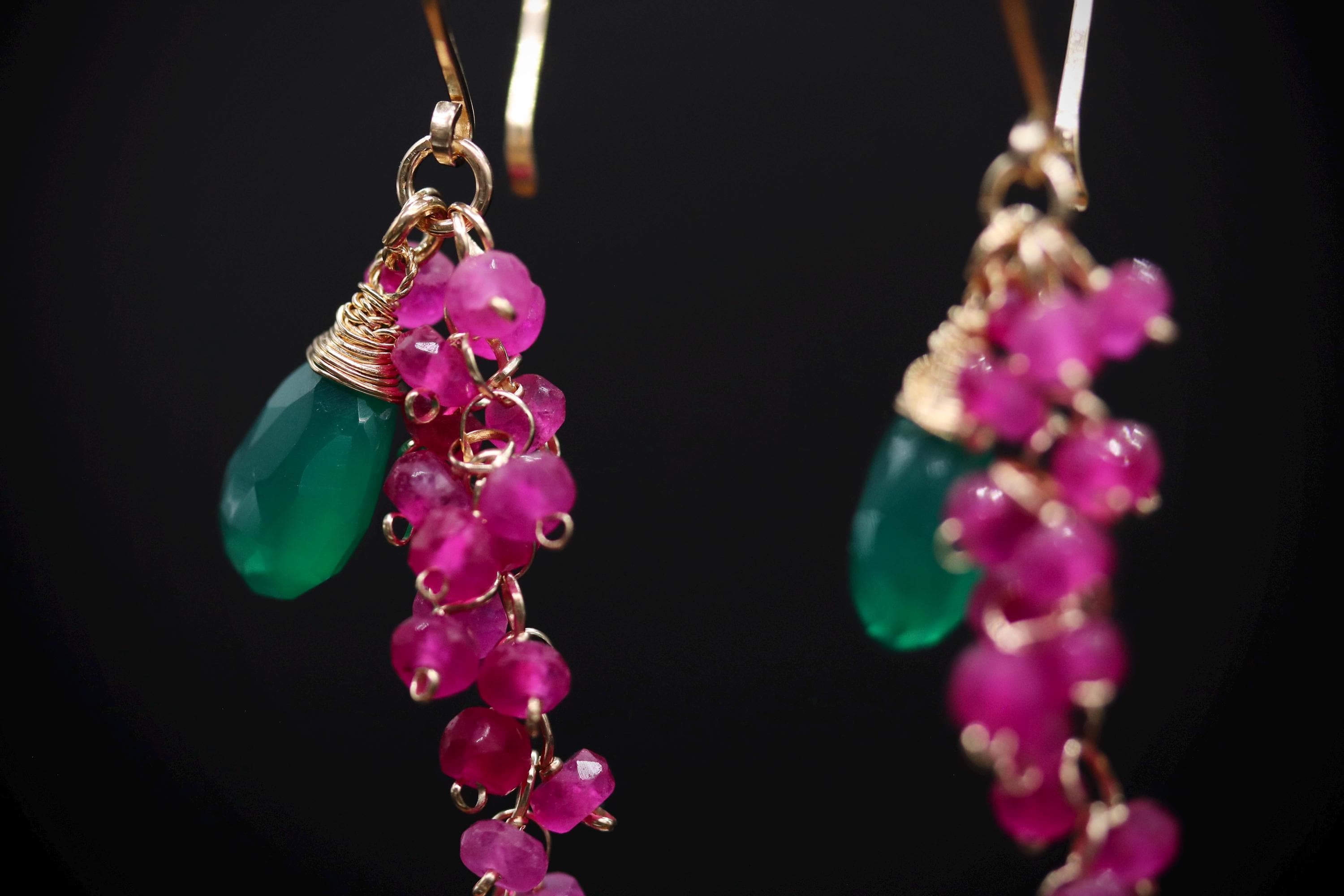 A close-up view of a pair of earrings with green and pink gemstones and dangling chains.