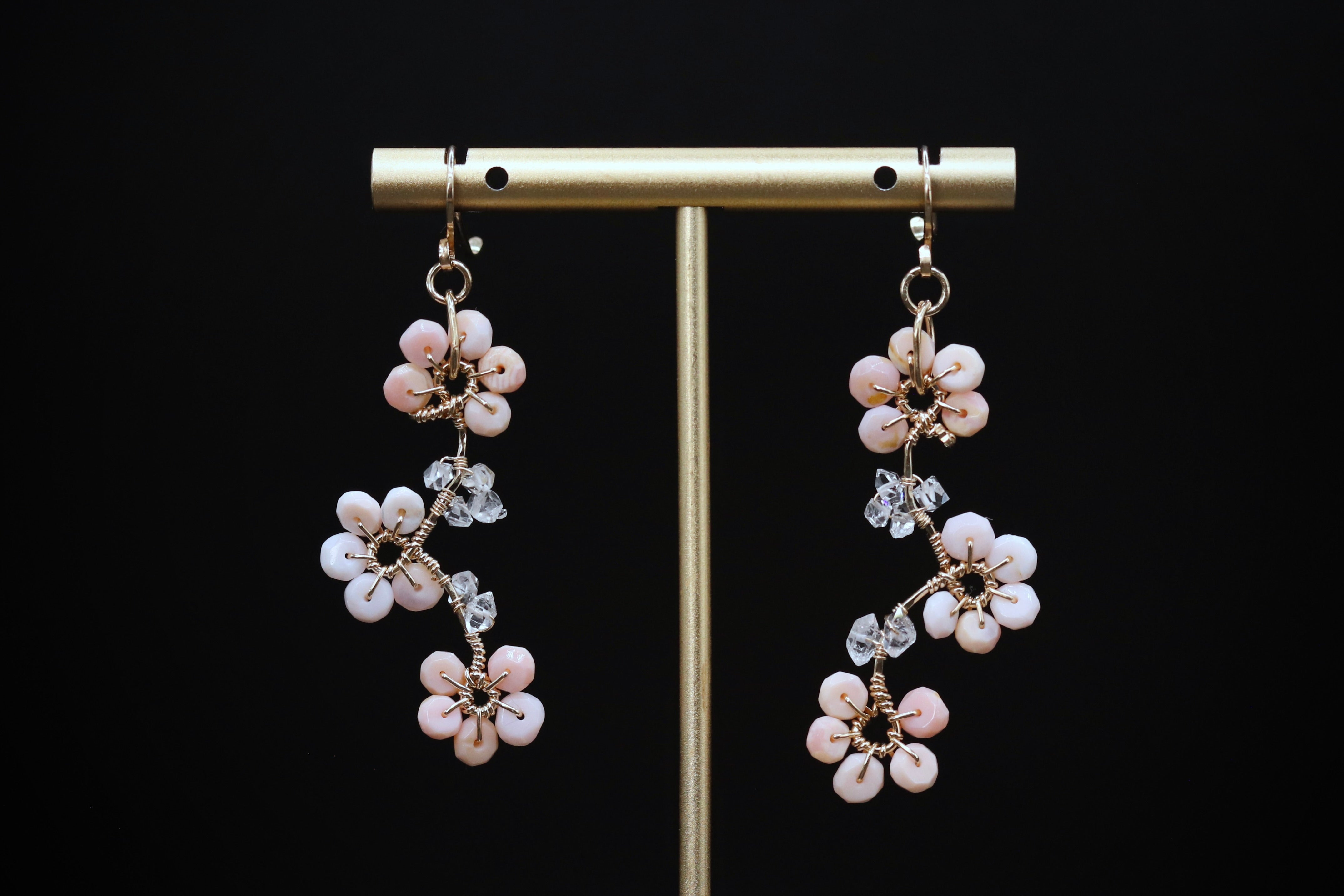 A pair of delicate earrings with white and pink flowers, hanging from a gold metal stand against a black background.