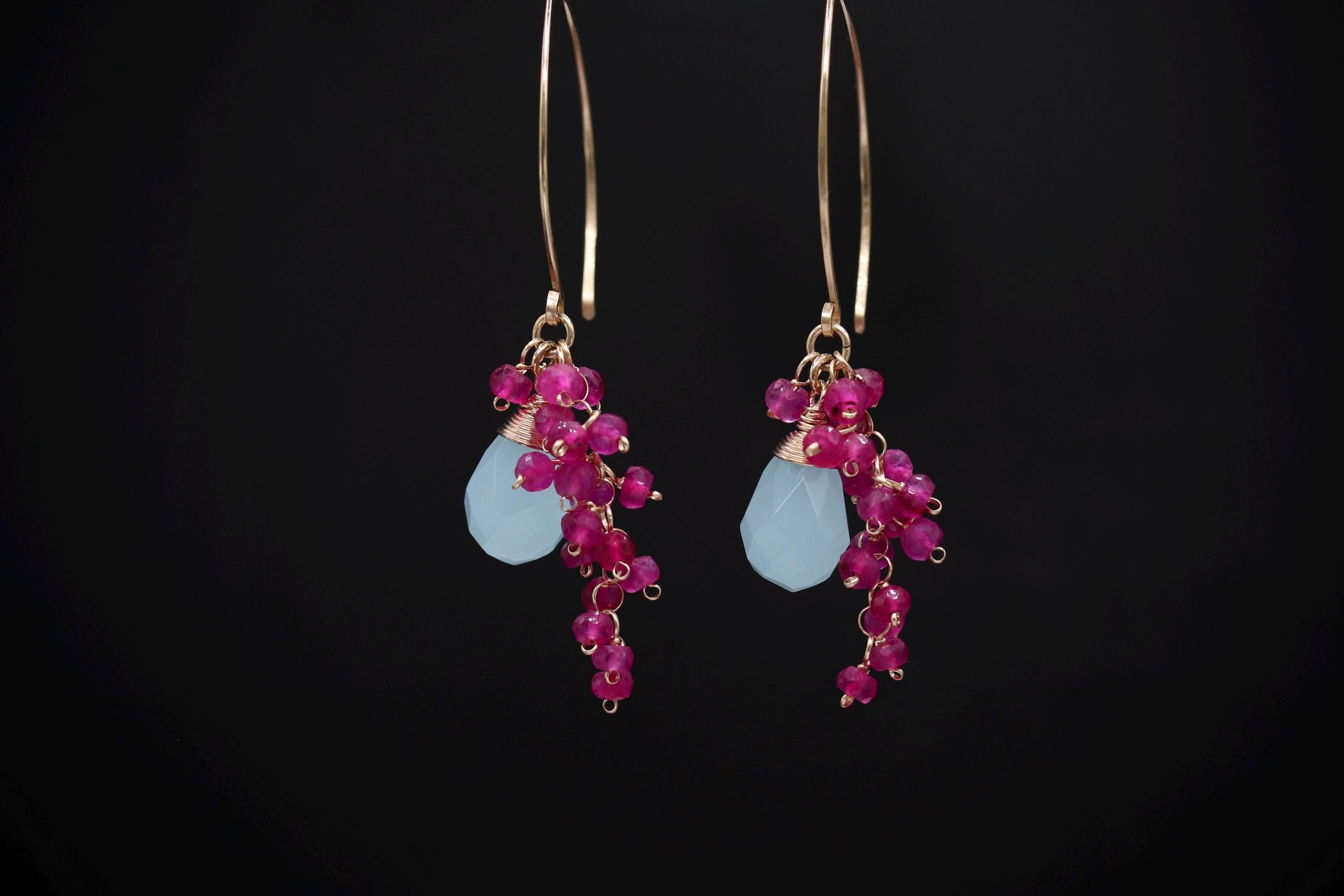 A pair of earrings with a combination of blue and pink gemstones, as well as small red gemstones, hanging from the ear hooks against a black background.