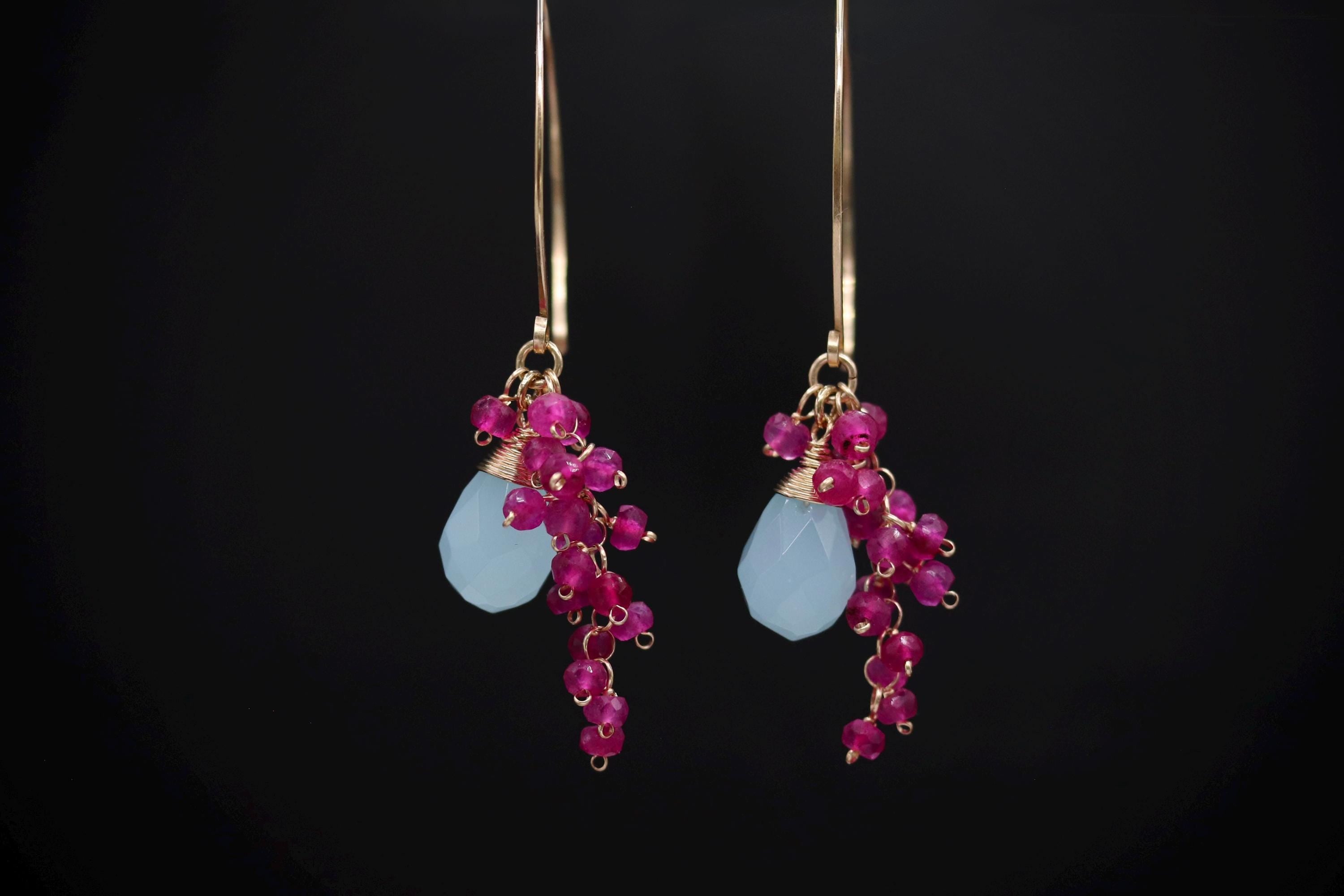A pair of elegant earrings with a combination of blue and pink gemstones, as well as small, dangling charms, set against a black background.