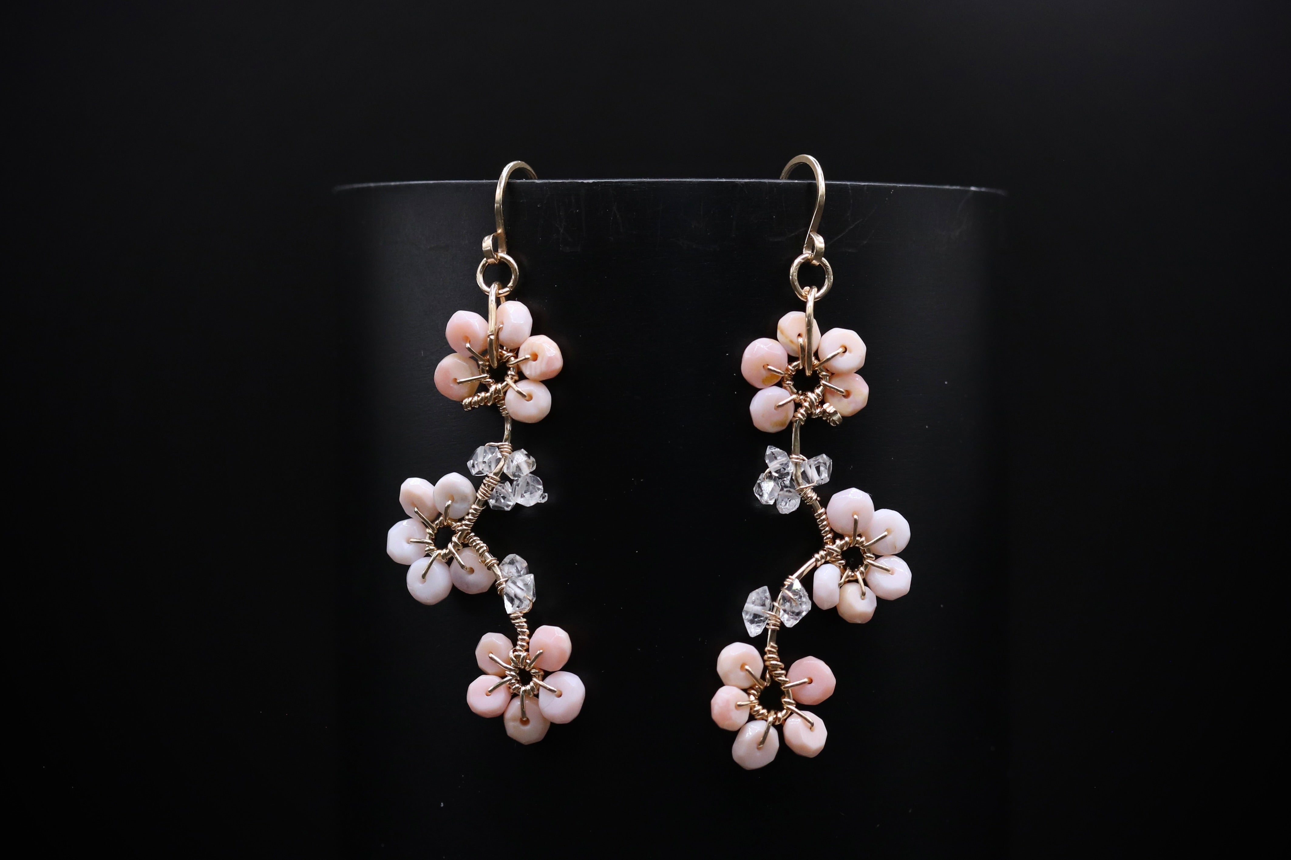 A pair of elegant earrings with delicate pink and white flower-shaped charms and crystal accents, displayed against a dark background.