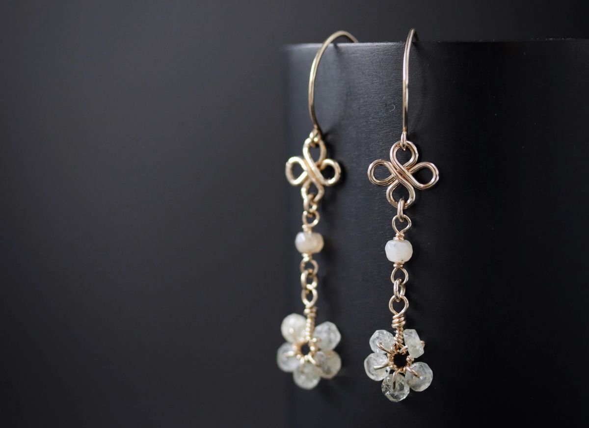 May include: Gold earrings with a delicate knot design and a cluster of white gemstones at the bottom. The earrings are hanging from a black surface.