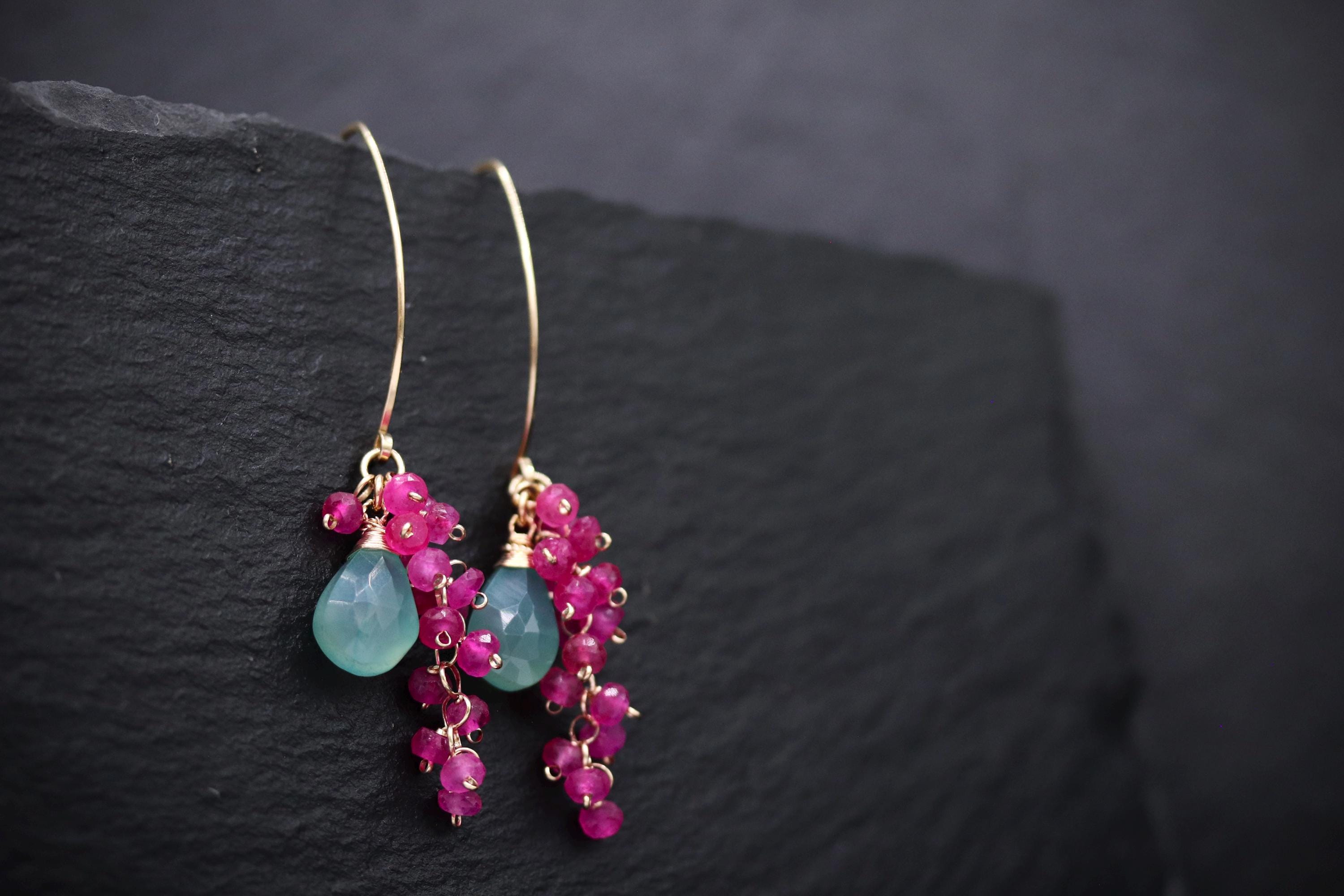 A pair of earrings with green and pink gemstones hanging from a gold hook against a dark background.