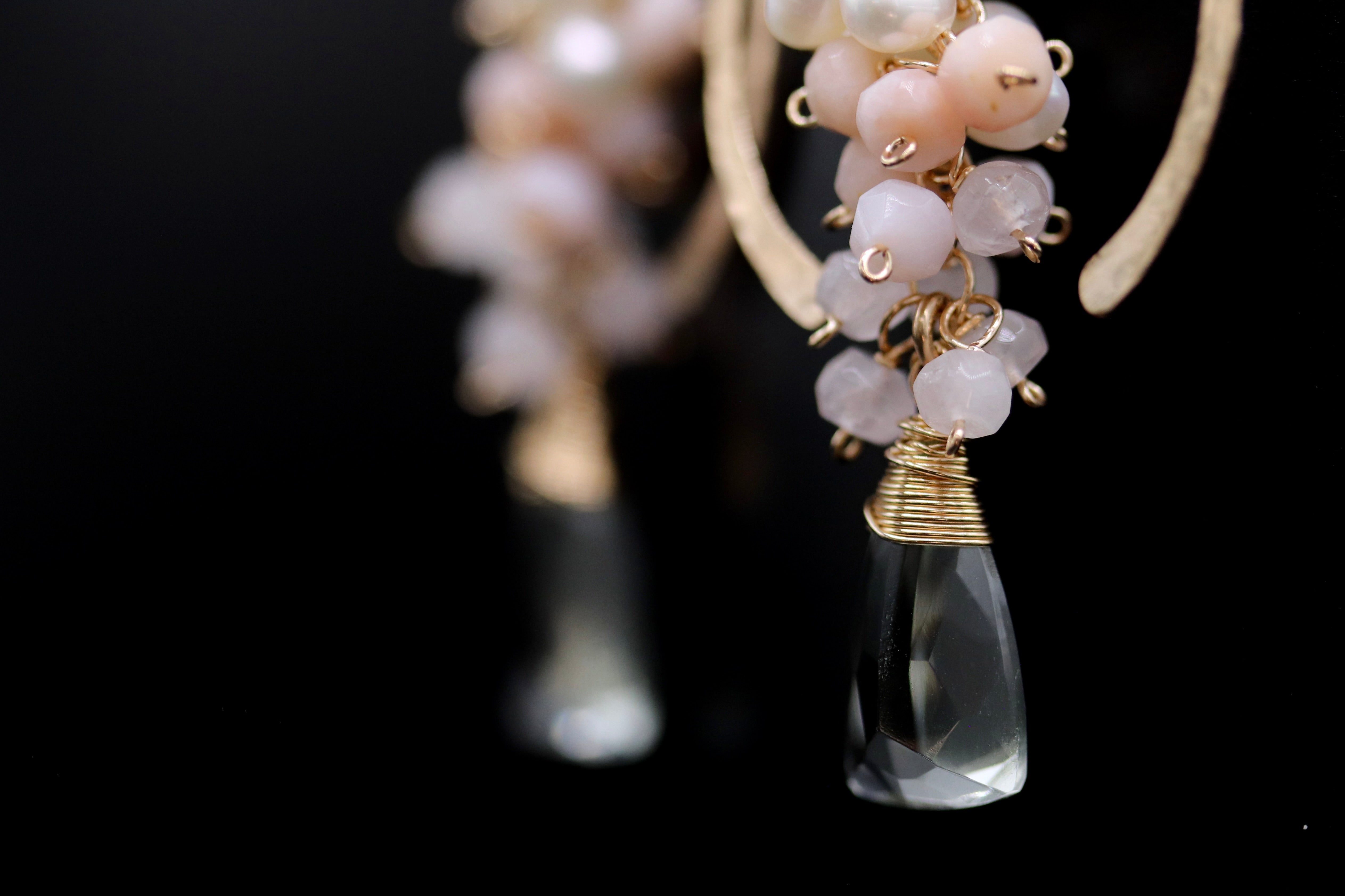 A close-up view of a pair of earrings with a clear crystal pendant and a cluster of small, round, pink and white stones or beads attached to the earring.