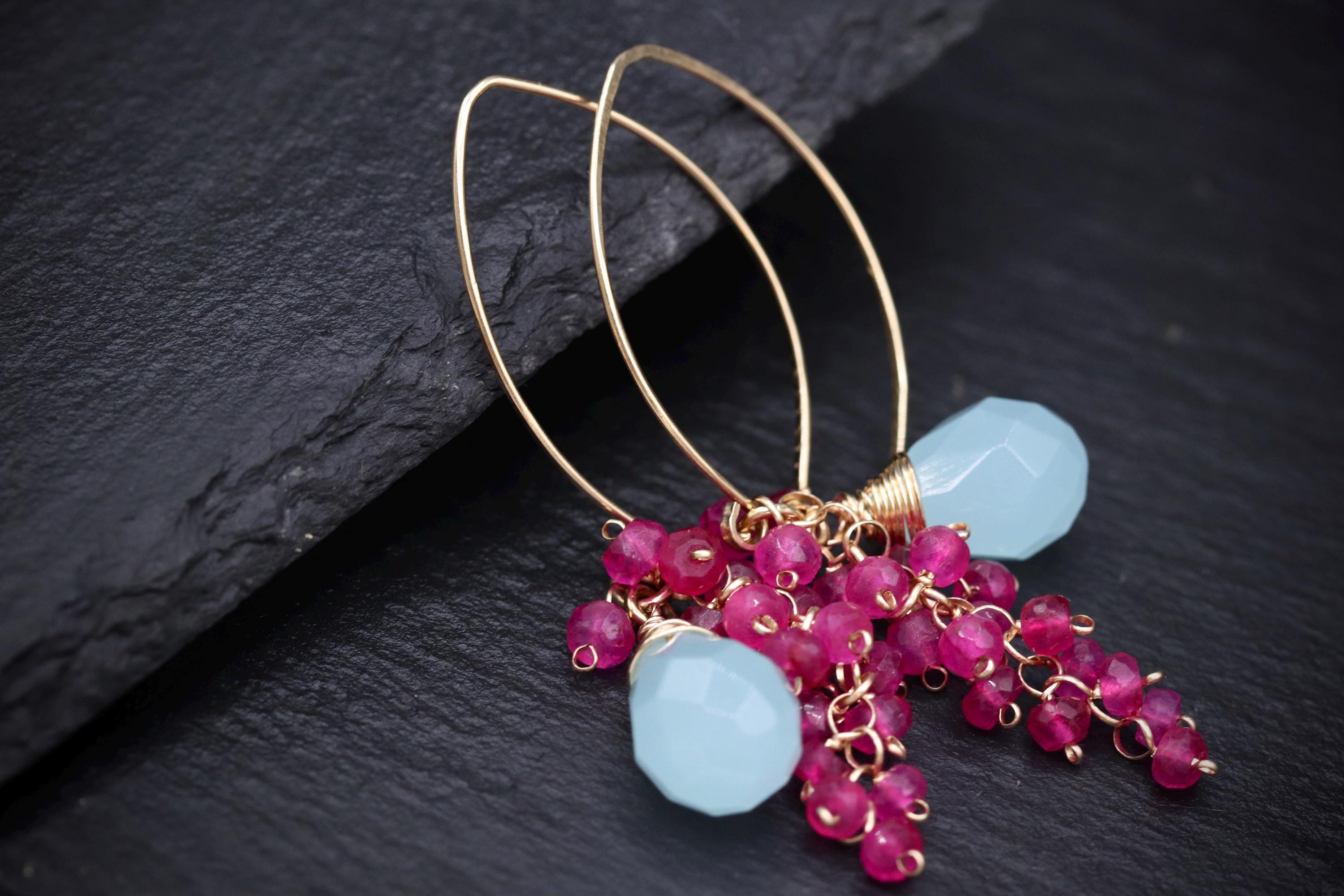 A pair of gold hoop earrings with blue and pink gemstone accents, resting on a dark surface.