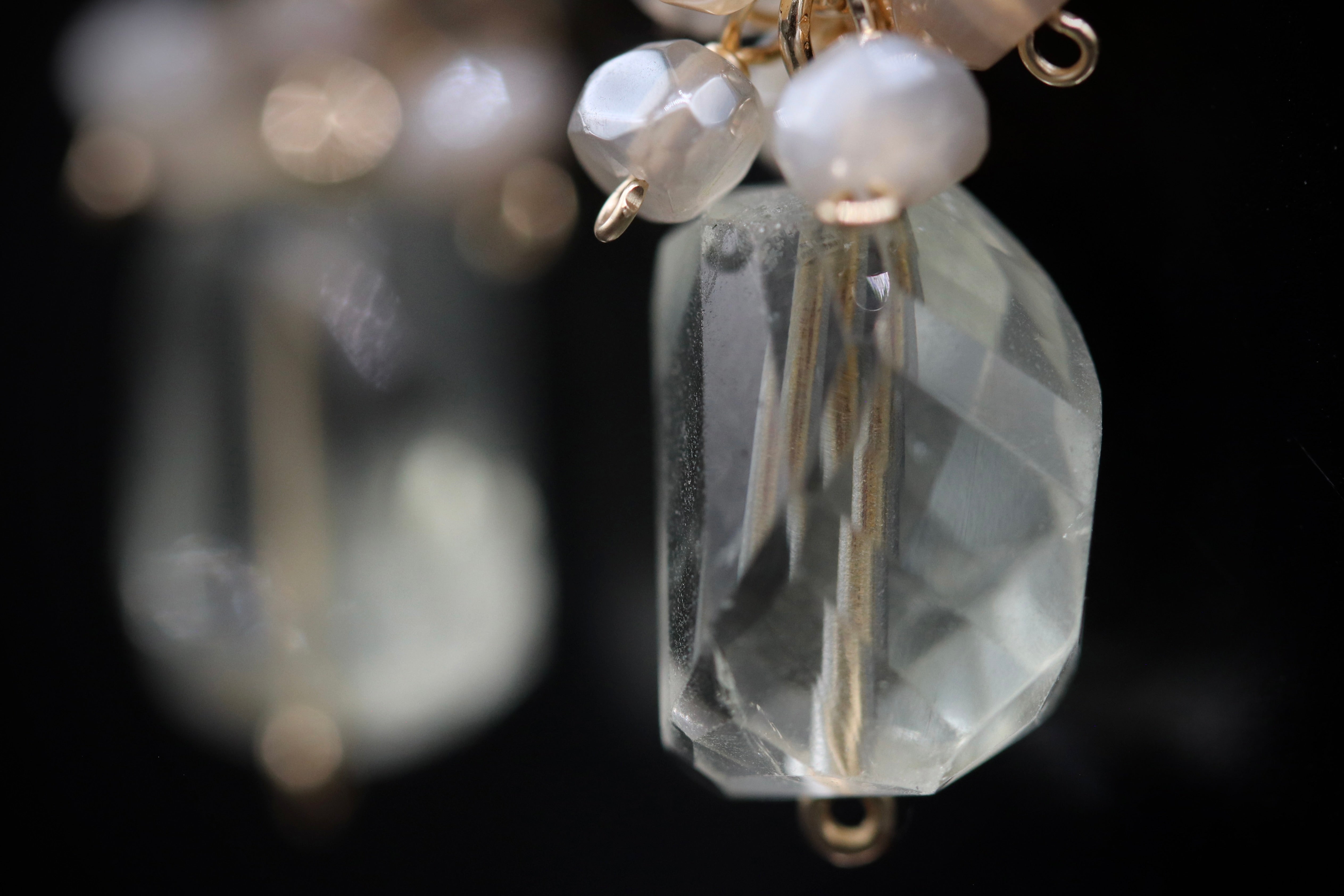 A close-up image of a clear crystal pendant with small white beads attached to it, set against a dark background.