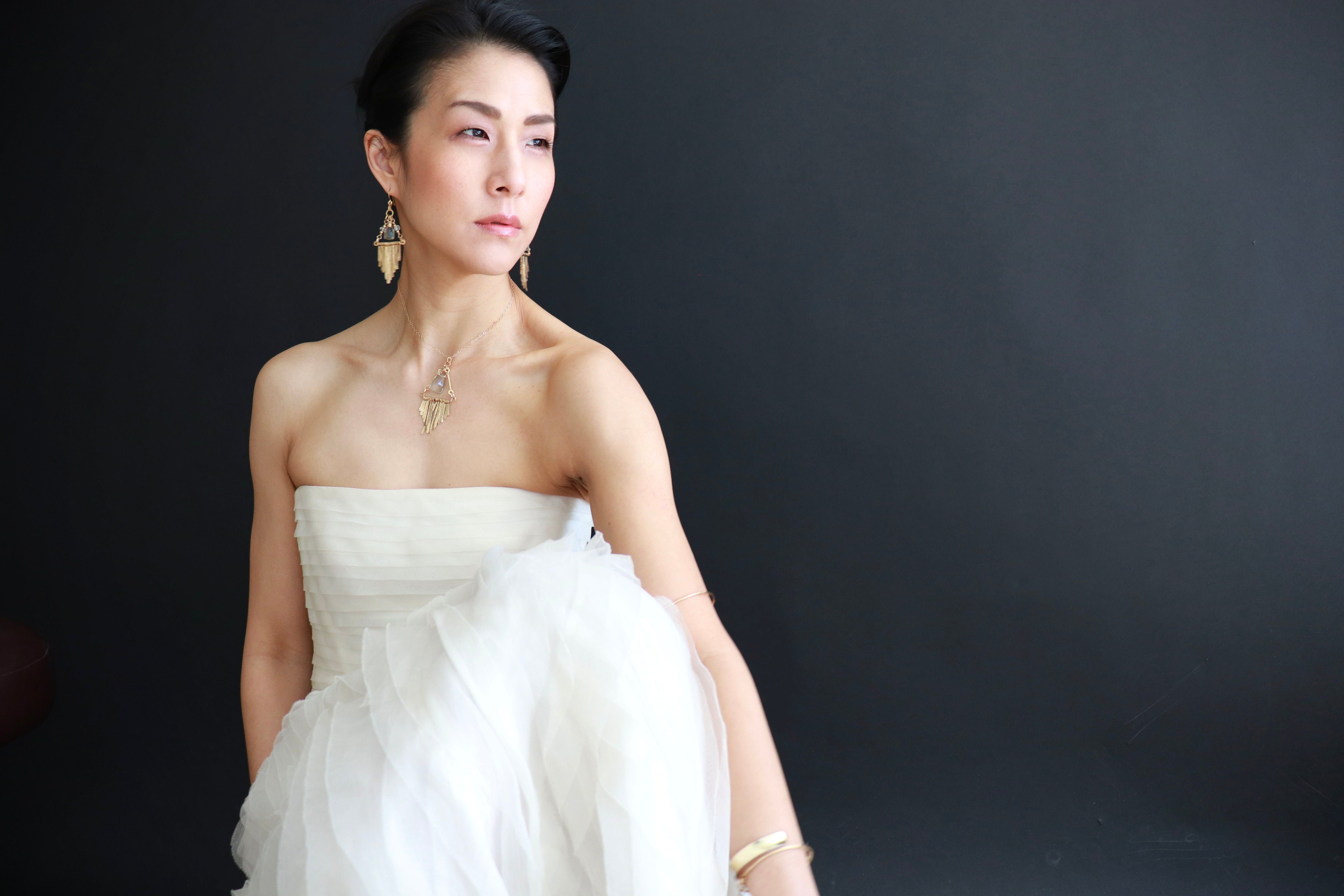 A woman in a white strapless dress poses against a dark background, wearing gold earrings and a necklace.