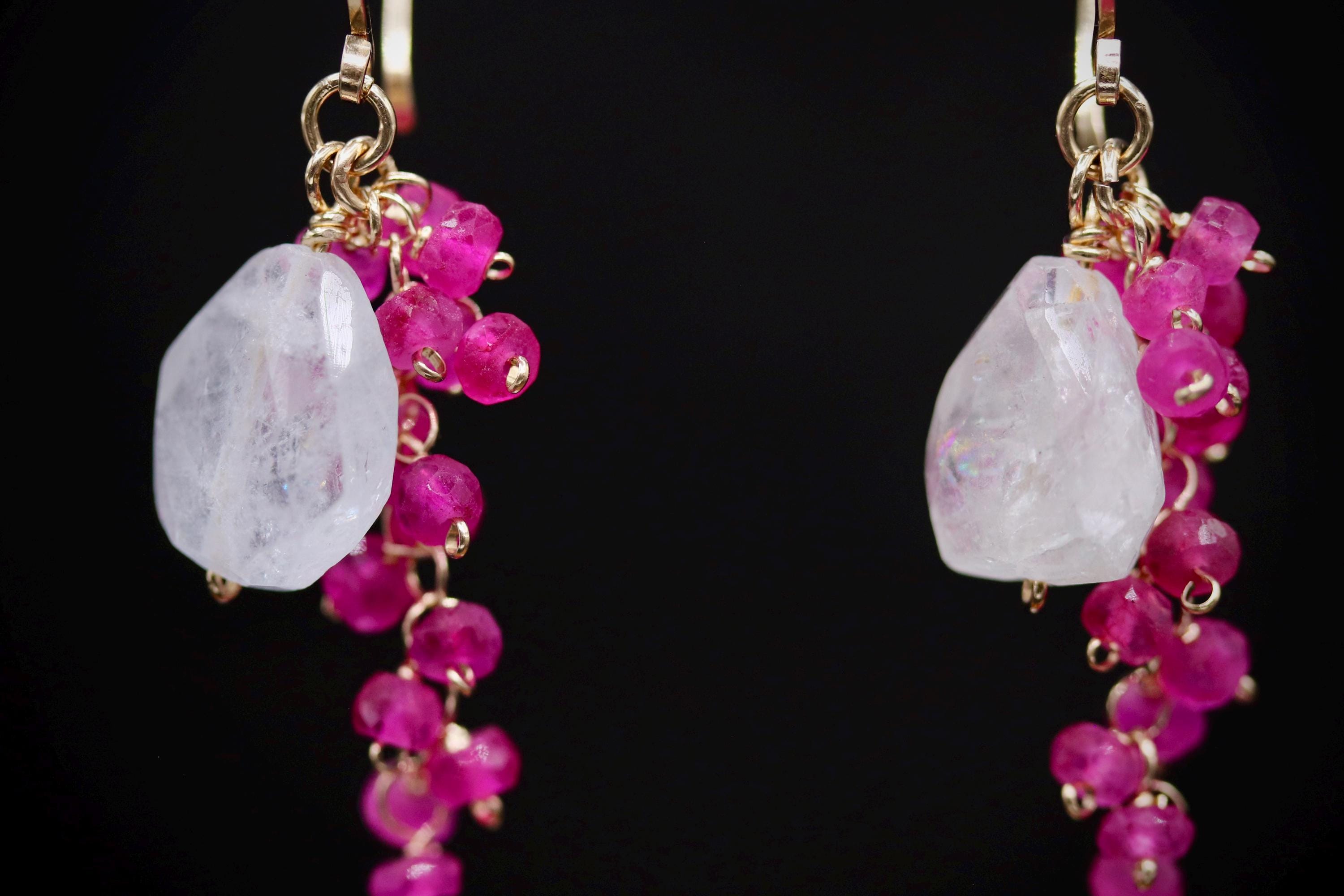 A pair of earrings with a combination of white and pink gemstones, as well as pink beads, hanging from gold hooks against a black background.