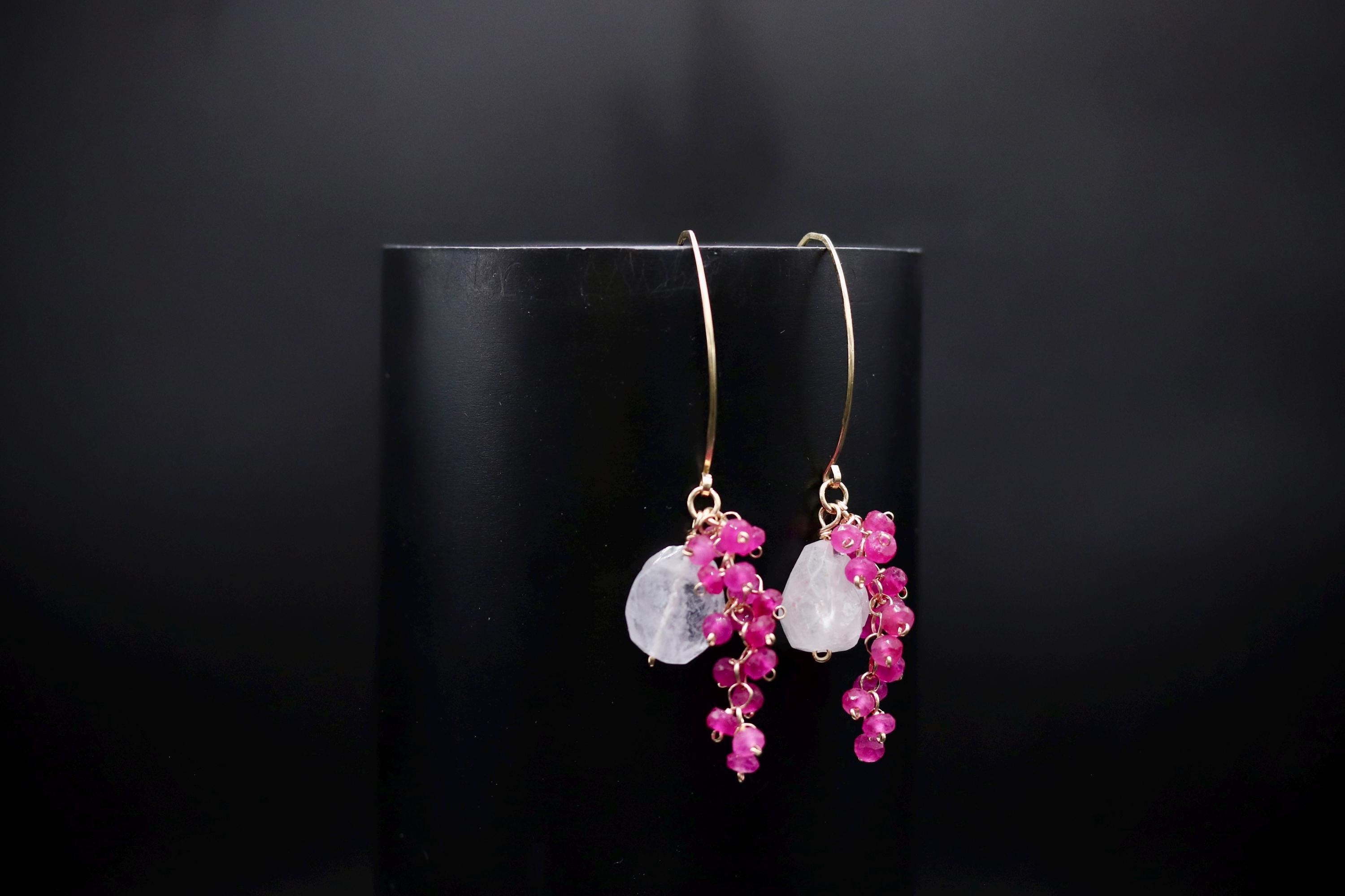 A pair of elegant earrings with a combination of pink and white stones, as well as small red stones, hanging from a gold hook against a black background.