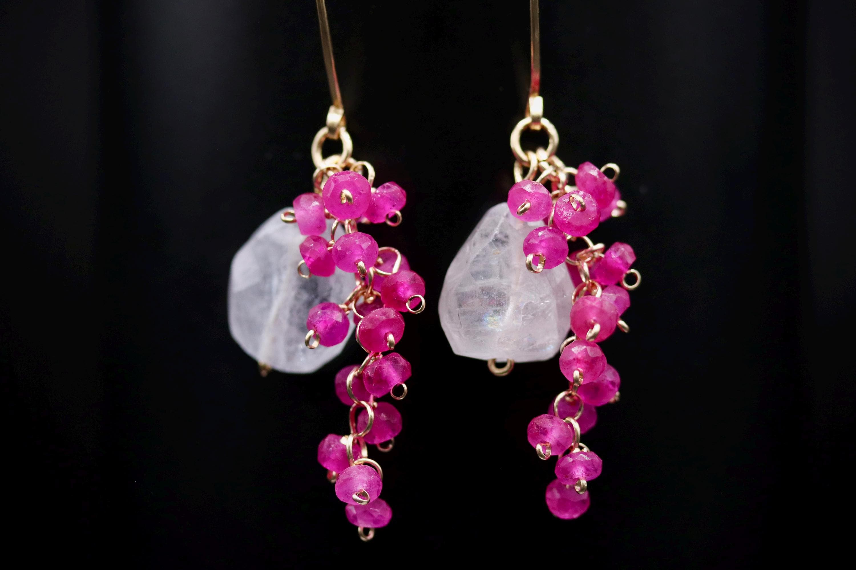A pair of earrings with clear gemstones and pink beads hanging from the ear hooks against a black background.
