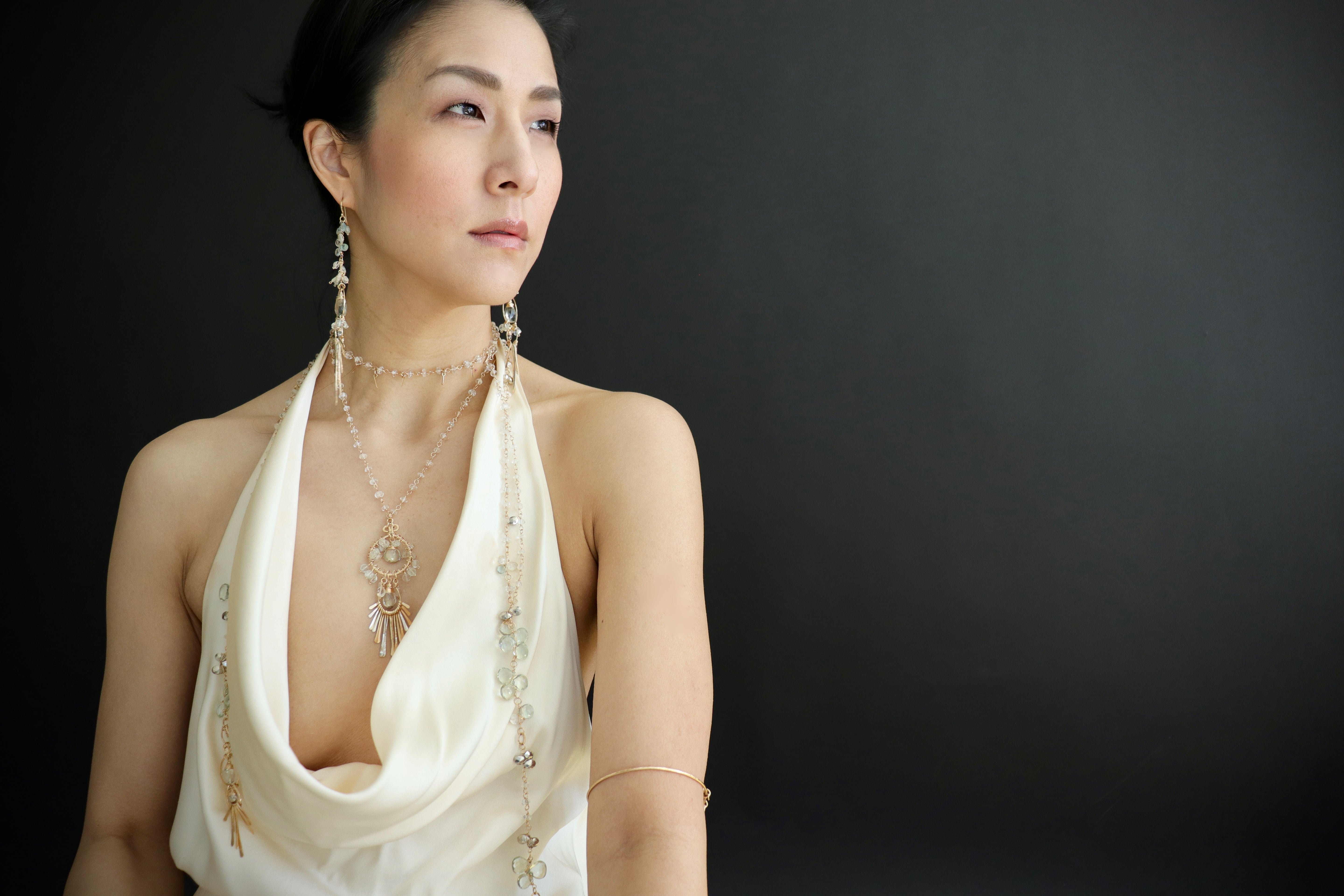 A woman wearing a white dress and earrings stands against a black background, looking off to the side with a thoughtful expression.