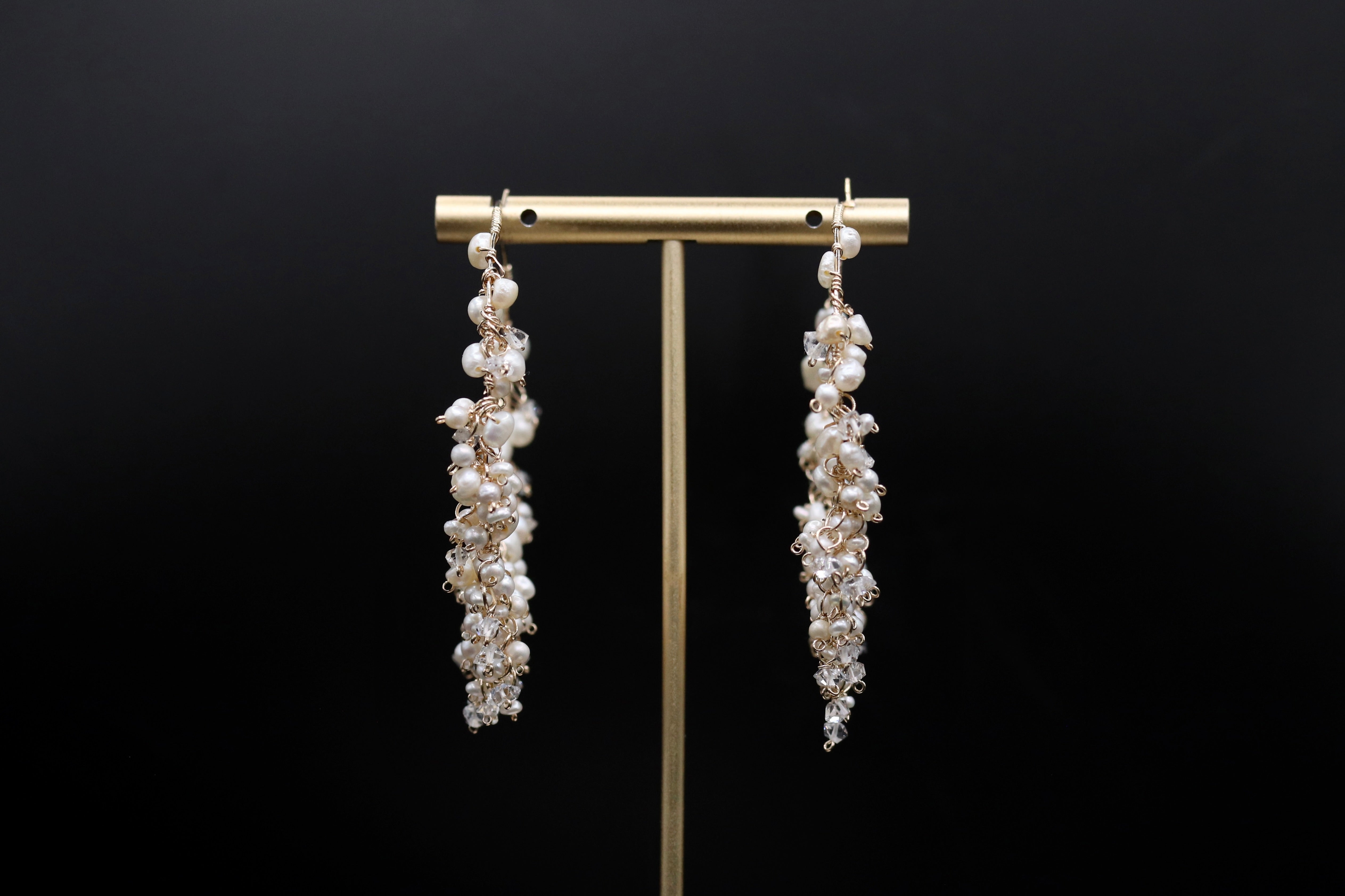 A pair of elegant earrings with long, dangling strands of pearls and crystals, displayed on a gold stand against a black background.