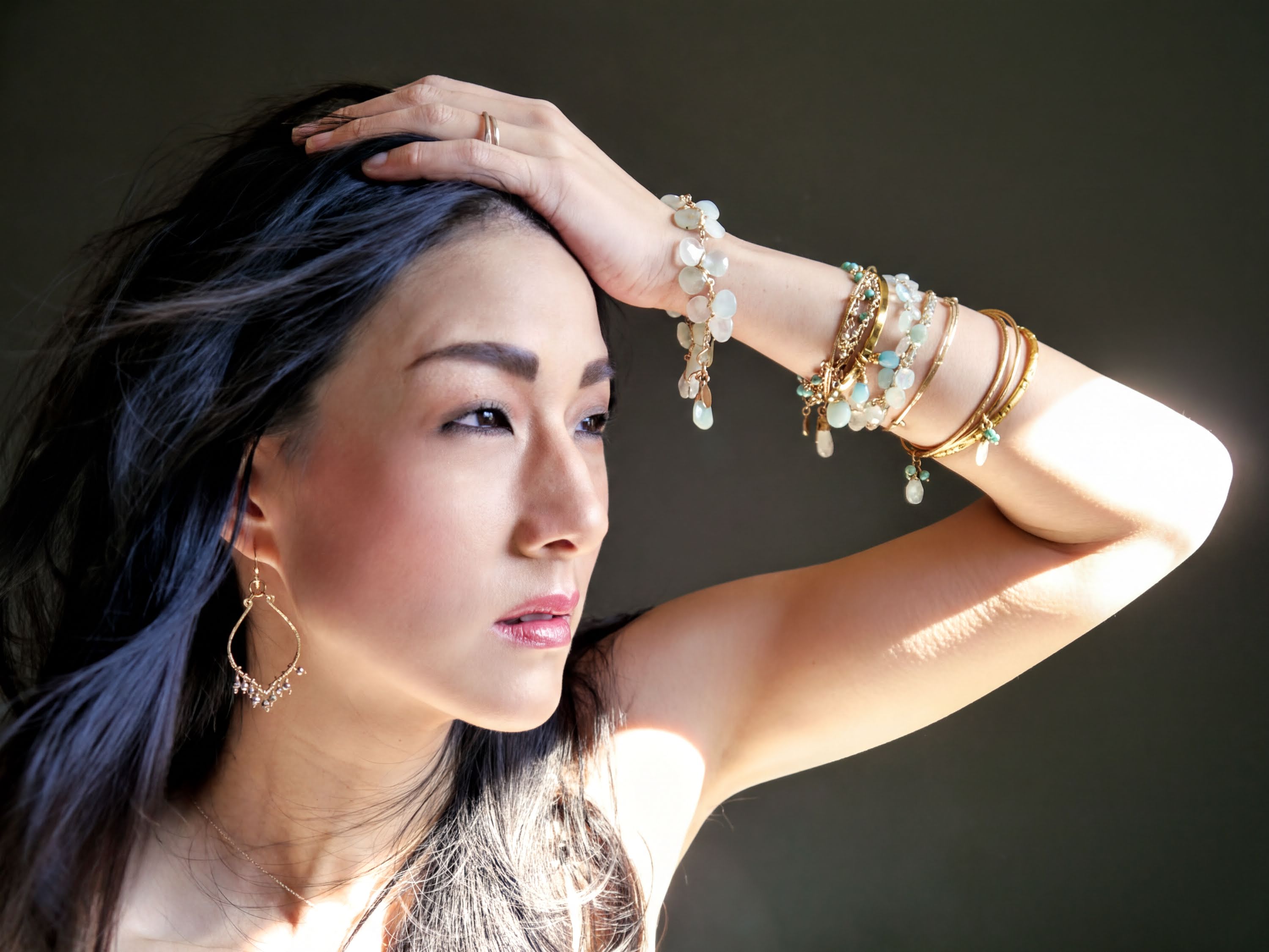 A woman with dark hair wearing multiple bracelets and earrings poses with her hand on her head, looking off to the side.
