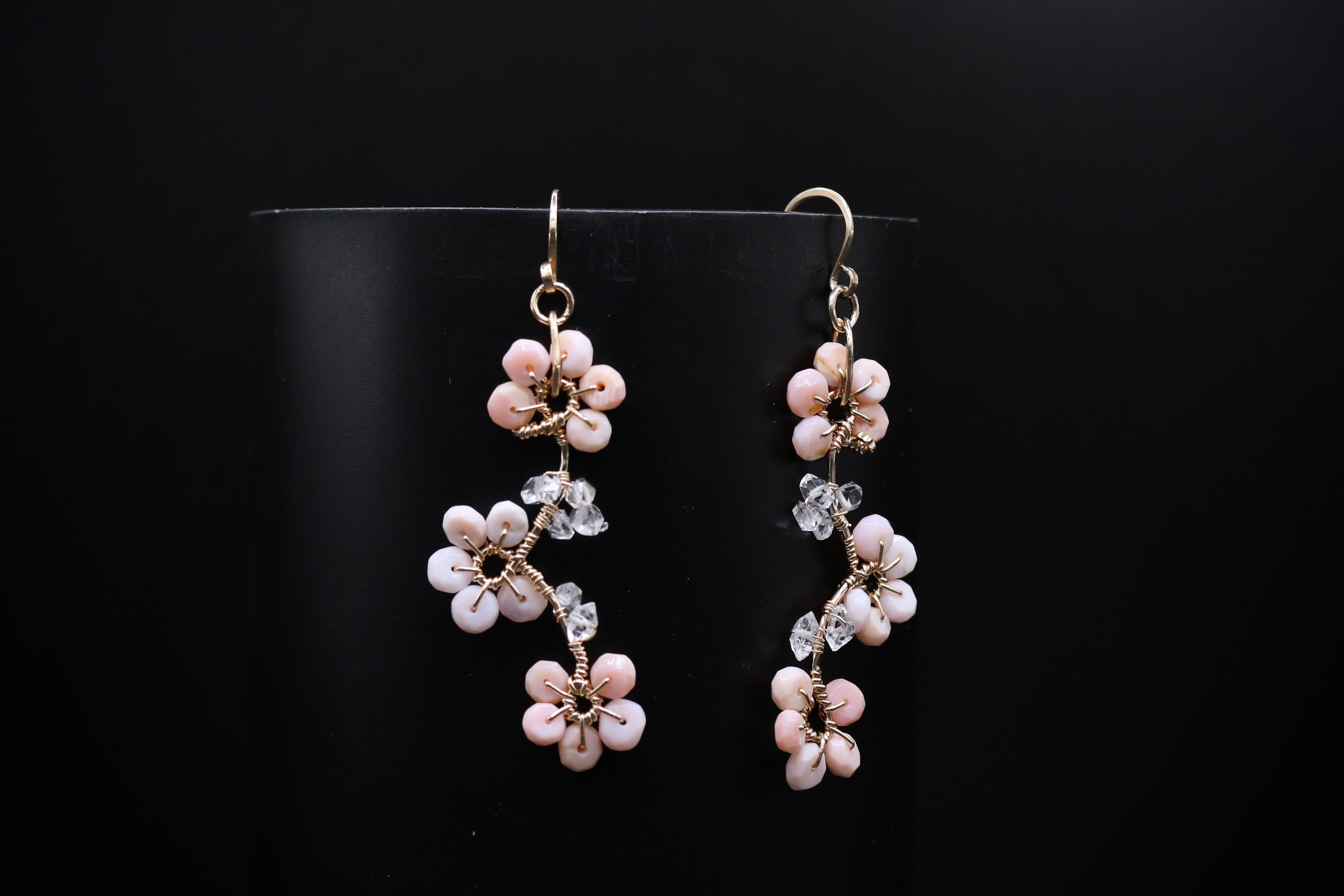 A pair of delicate earrings with small white and pink flowers and clear gemstones, displayed against a black background.