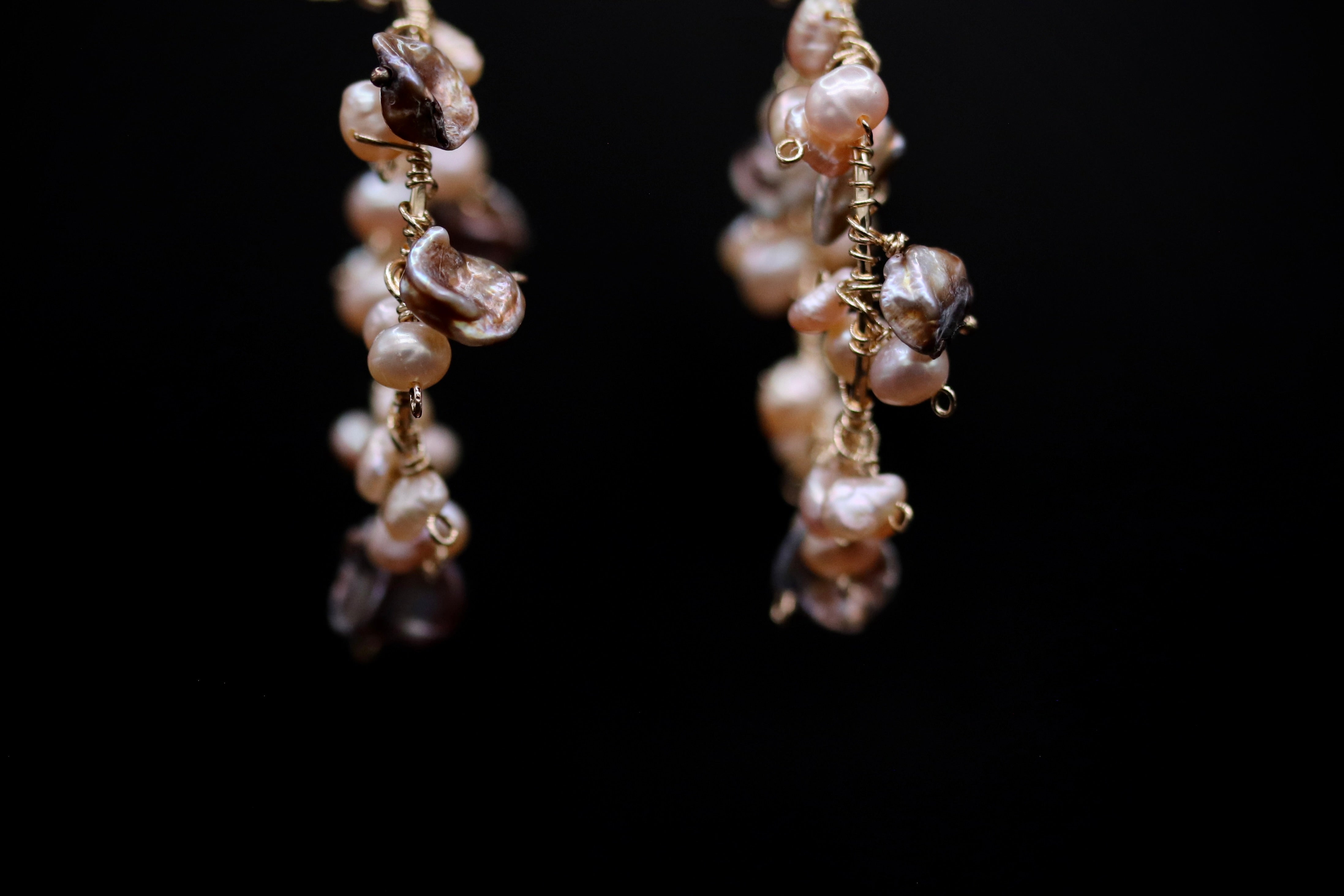 A pair of earrings with a combination of white and brown pearls and small, dangling charms against a black background.
