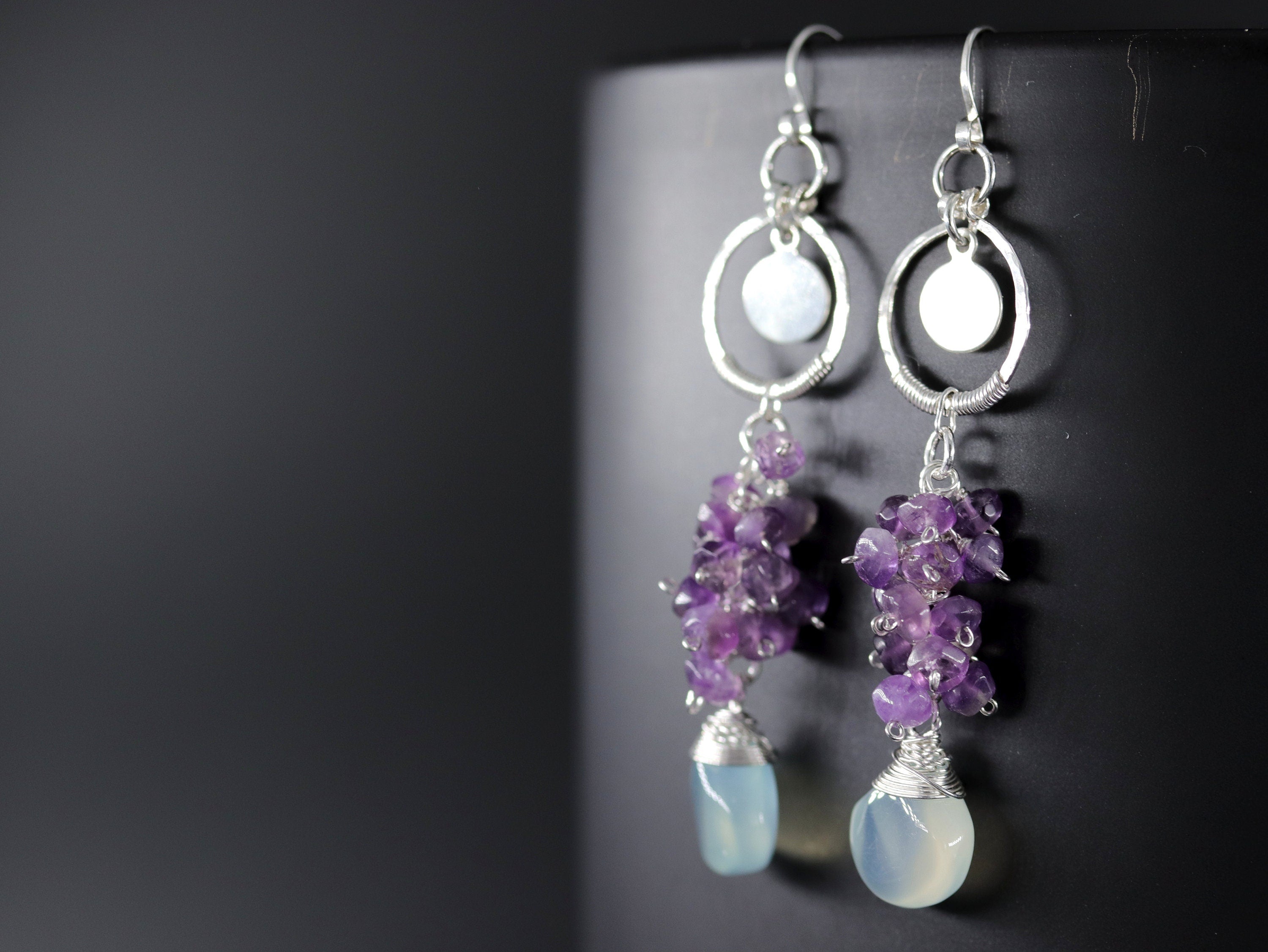 May include: Silver dangle earrings with a circle charm and a cluster of purple amethyst stones hanging from a teardrop shaped light blue chalcedony stone.