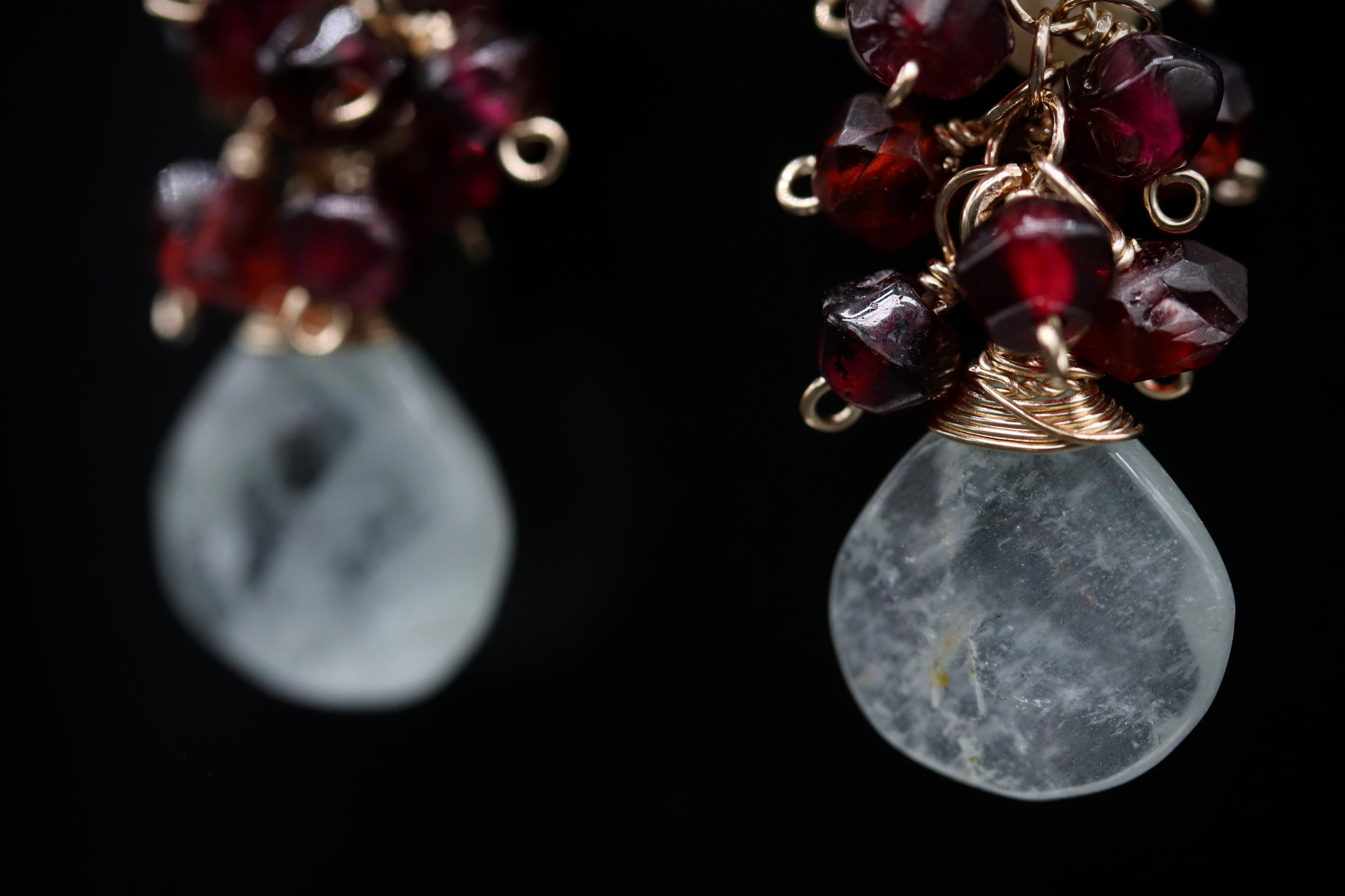 A close-up view of a pair of earrings with a combination of clear and maroon gemstones, as well as a metallic wire and bead design.