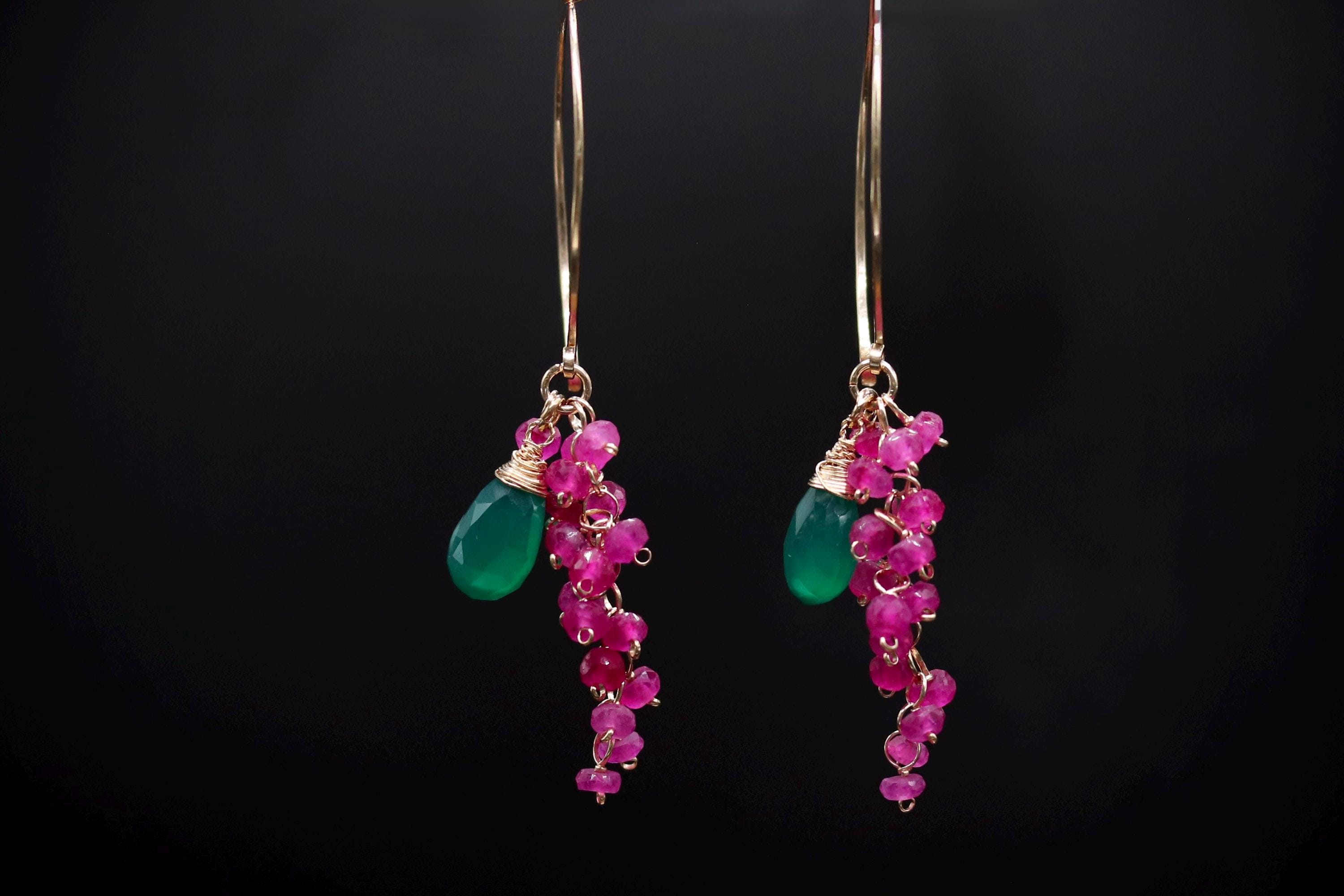 A pair of earrings with green gemstones and pink gemstones hanging from the ear hooks against a black background.