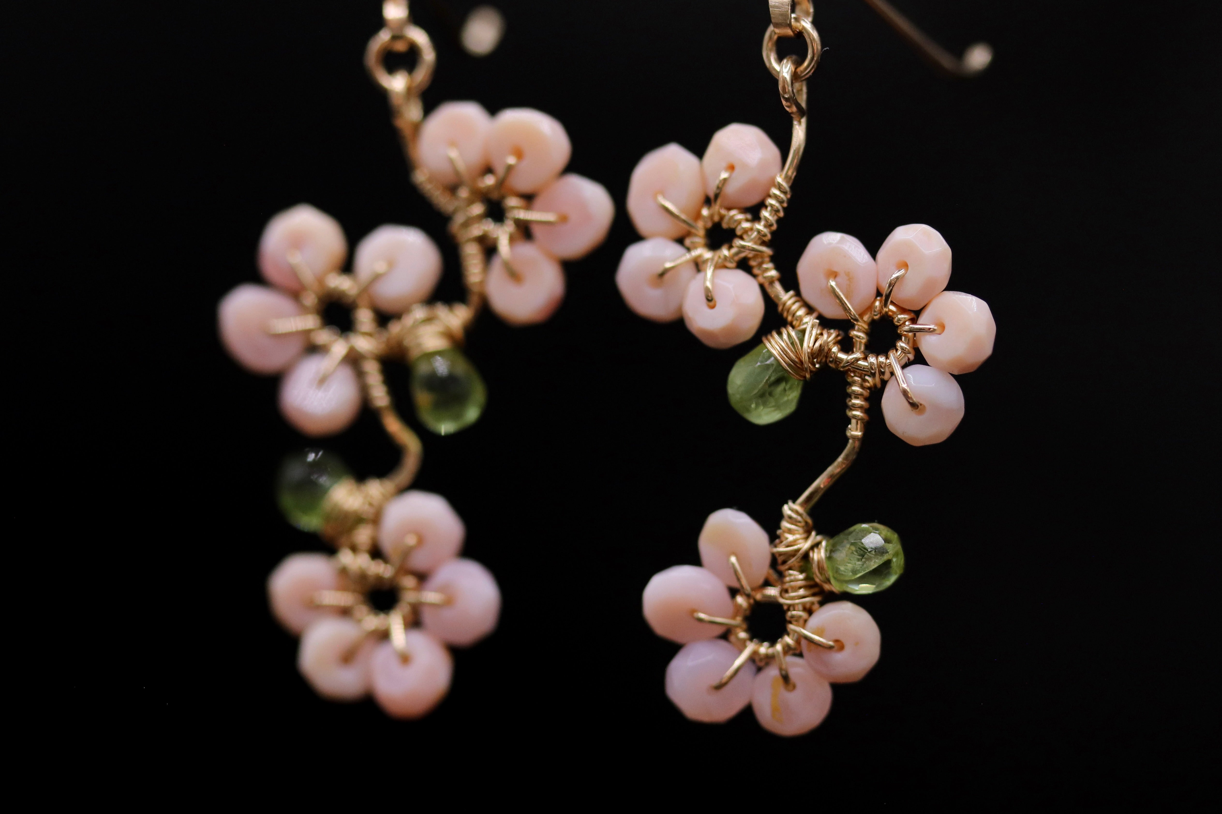 A pair of delicate earrings with pink flowers and green stones, suspended from a gold chain against a black background.