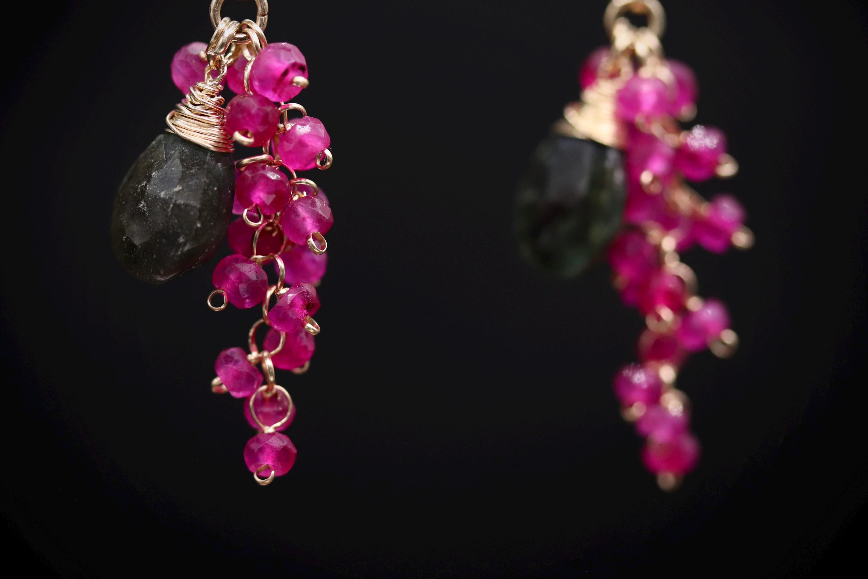 A close-up view of a pair of earrings with a combination of red and black gemstones, as well as dangling beads in a dark background.