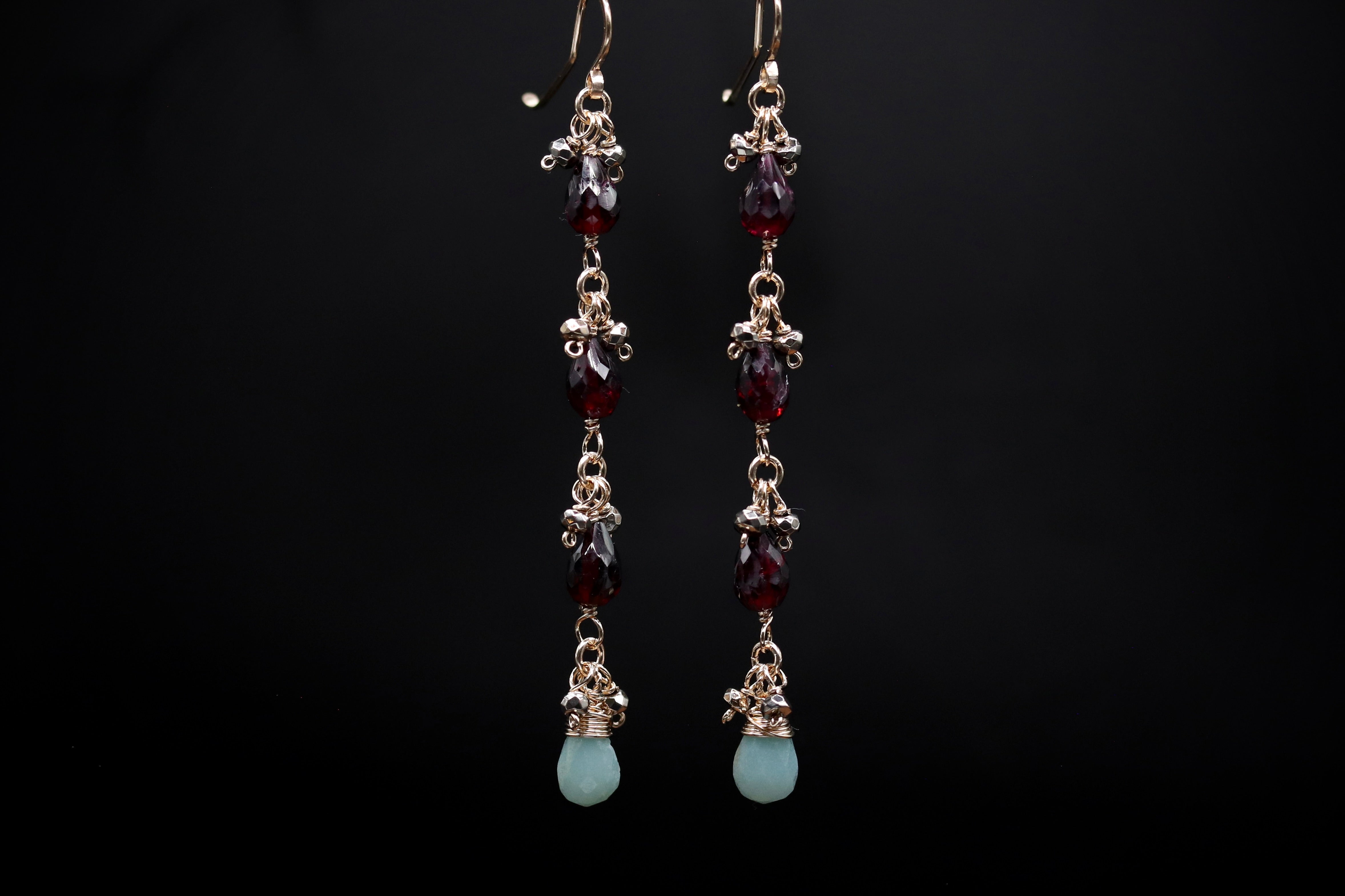 A pair of elegant earrings with a row of small, dangling gemstones in shades of red and green, set in a metallic frame against a dark background.