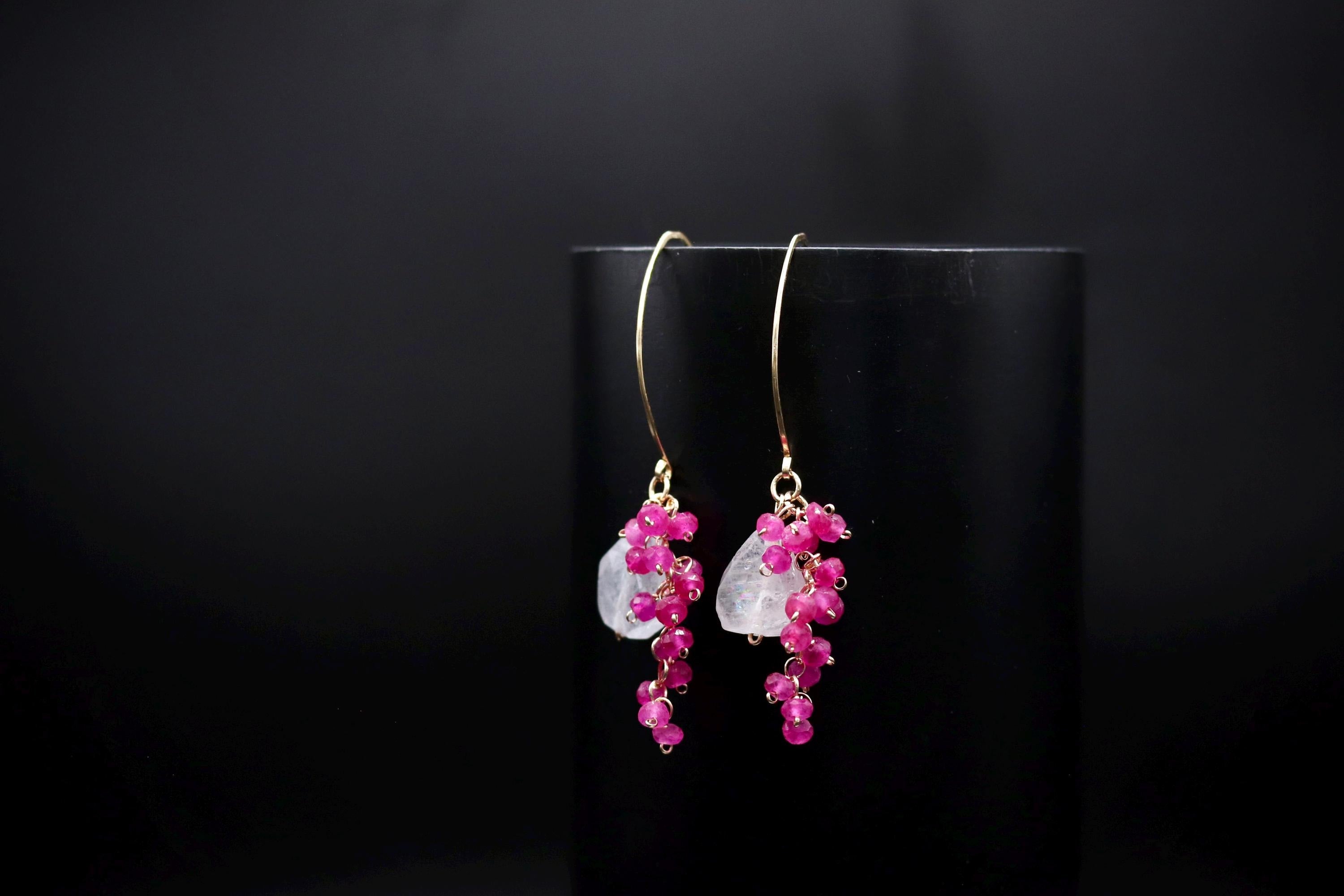 A pair of elegant earrings with a combination of pink gemstones and white stones, hanging from a gold hook against a black background.