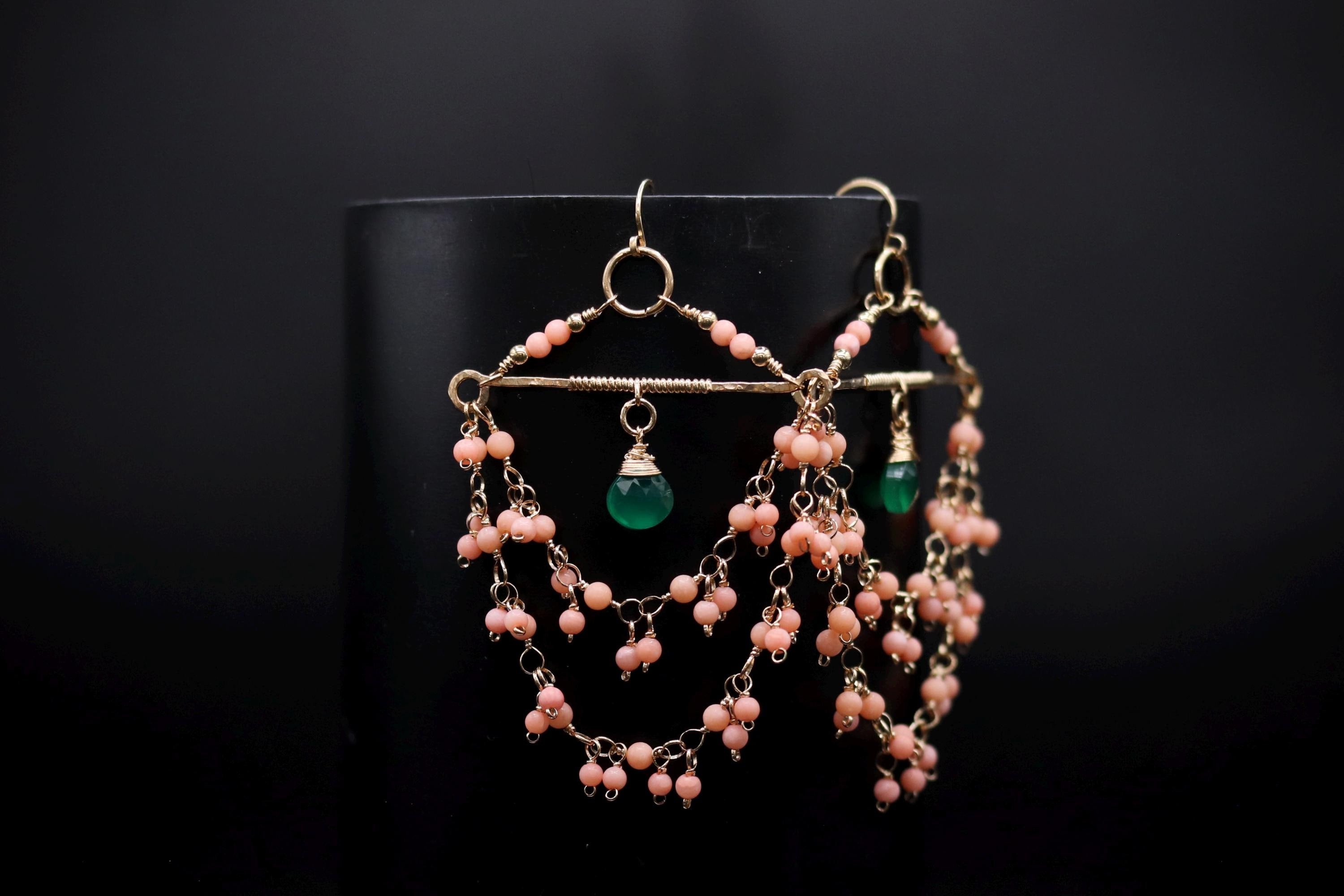 A pair of earrings with a black background and a gold frame, featuring a green gemstone and pink beads.