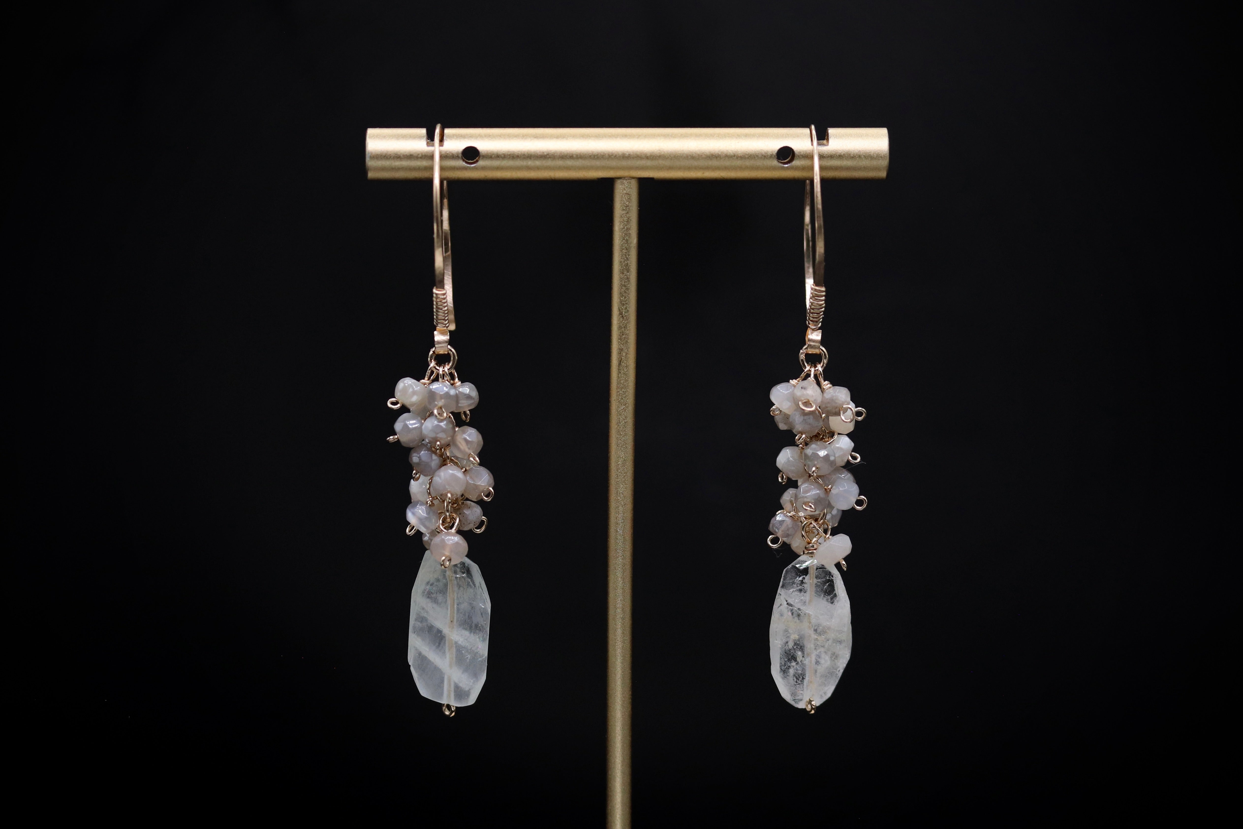 A pair of elegant earrings with clear gemstone pendants and dangling metal hooks, displayed on a gold stand against a black background.