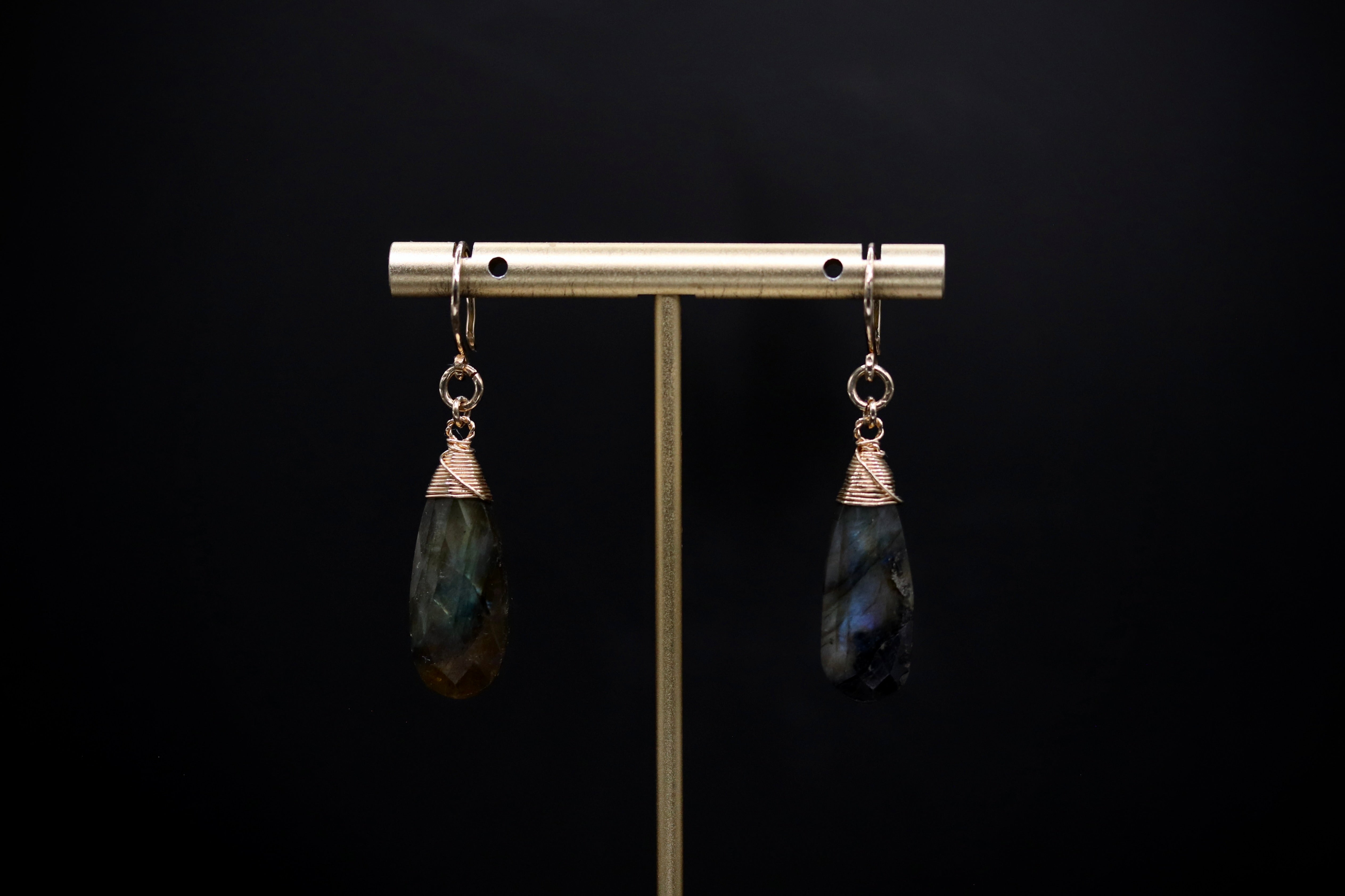 A pair of earrings with dark blue and green stones hanging from a gold-colored metal stand against a black background.