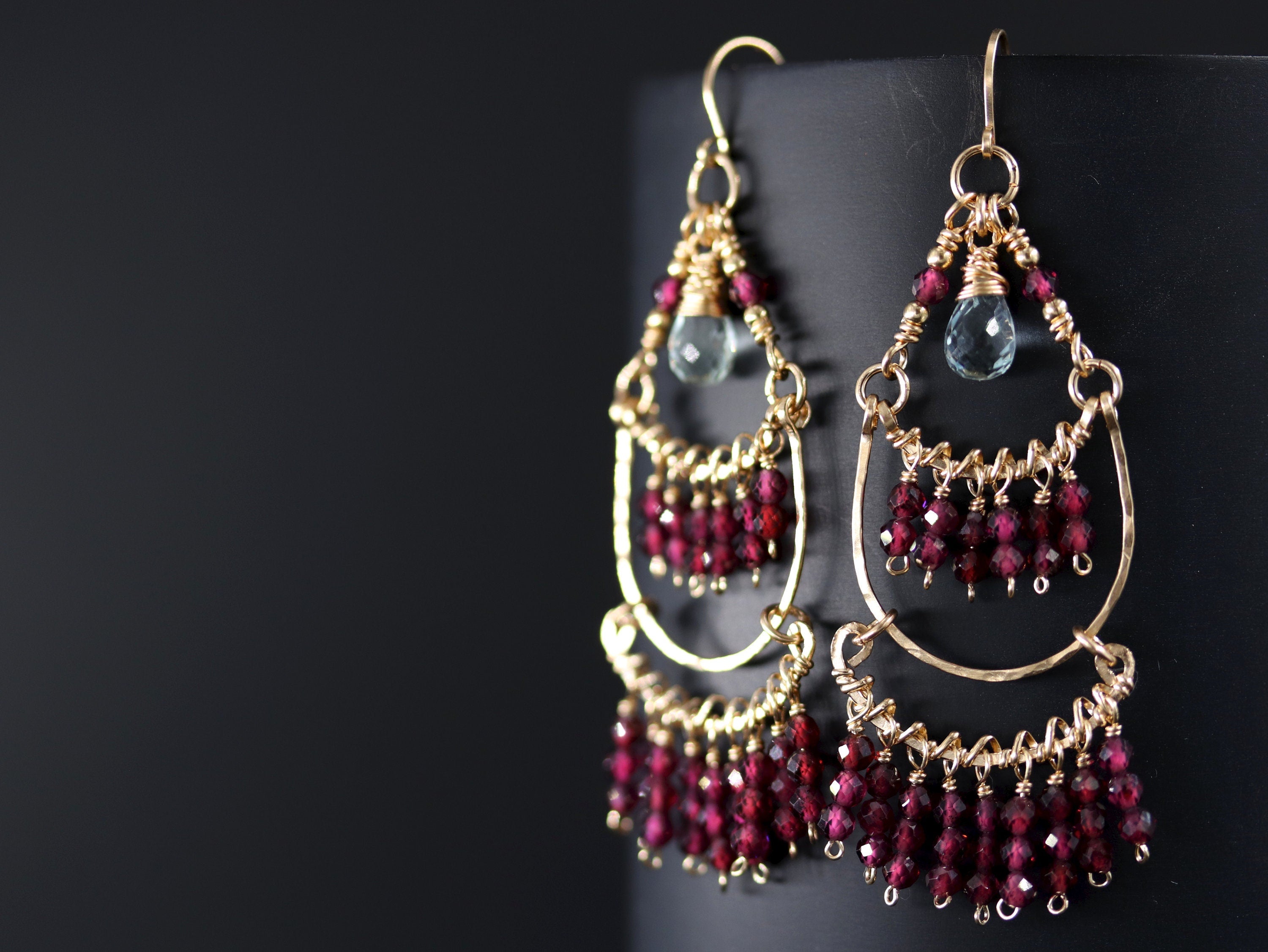 May include: Gold earrings with a teardrop-shaped aquamarine gemstone and dangling red garnet beads.