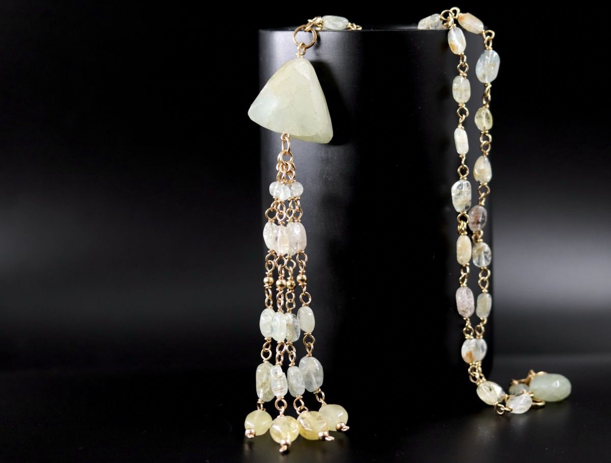May include: A long gold chain necklace with a large, triangular, light green gemstone pendant. The pendant is attached to a tassel of smaller, light green gemstones and gold beads.