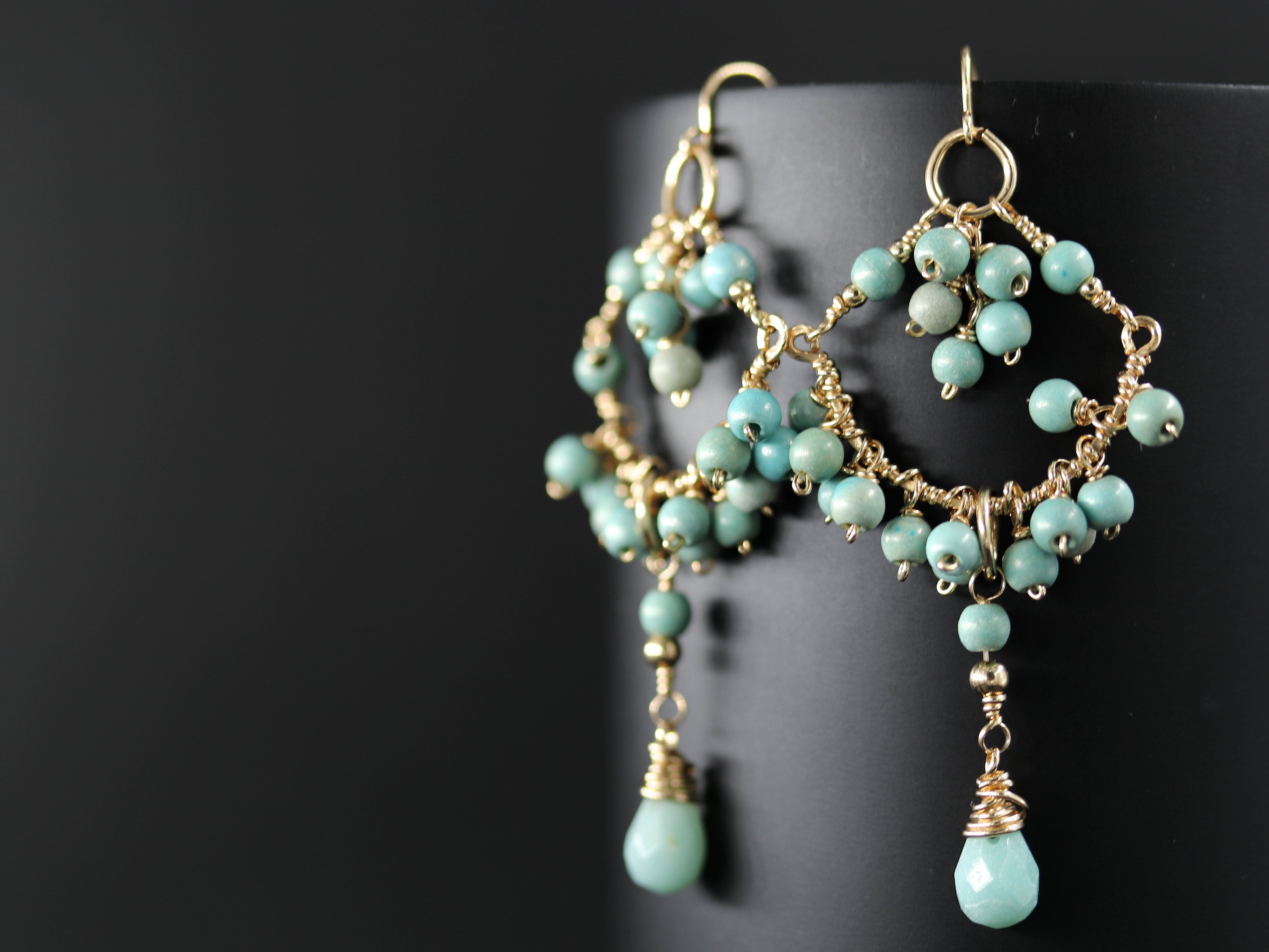 May include: Gold-toned chandelier earrings with turquoise-colored beads. The earrings feature a delicate wire design with multiple tiers of small, round beads and teardrop-shaped dangles. The earrings are displayed against a dark background.