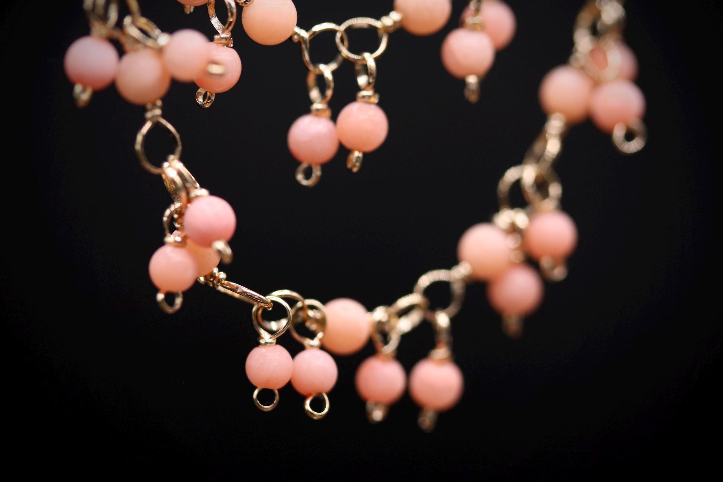 A close-up view of a delicate necklace with numerous small, round, pink beads hanging from a chain against a dark background.