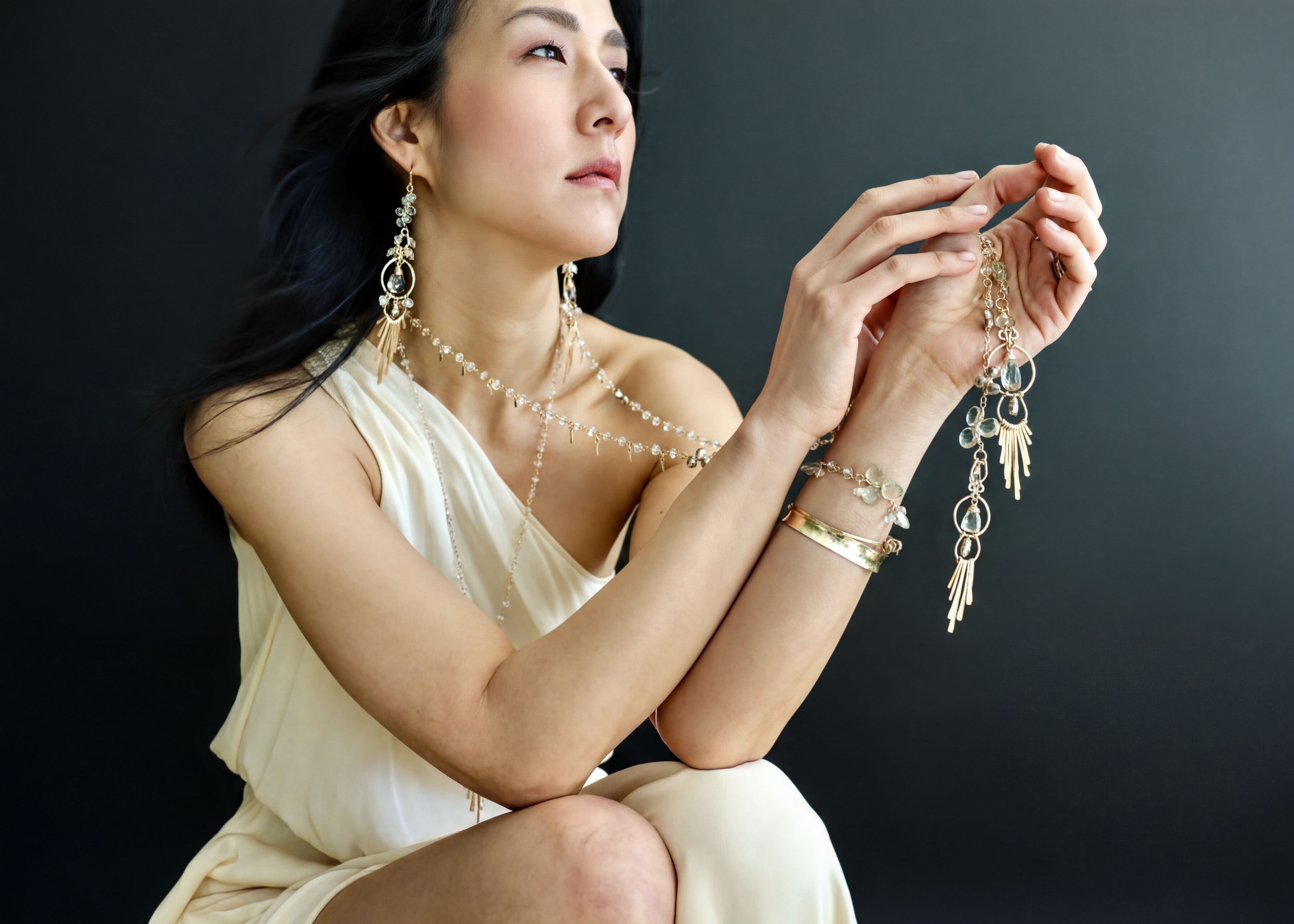 A woman in a white dress is holding a necklace and a bracelet, with her hands clasped together.