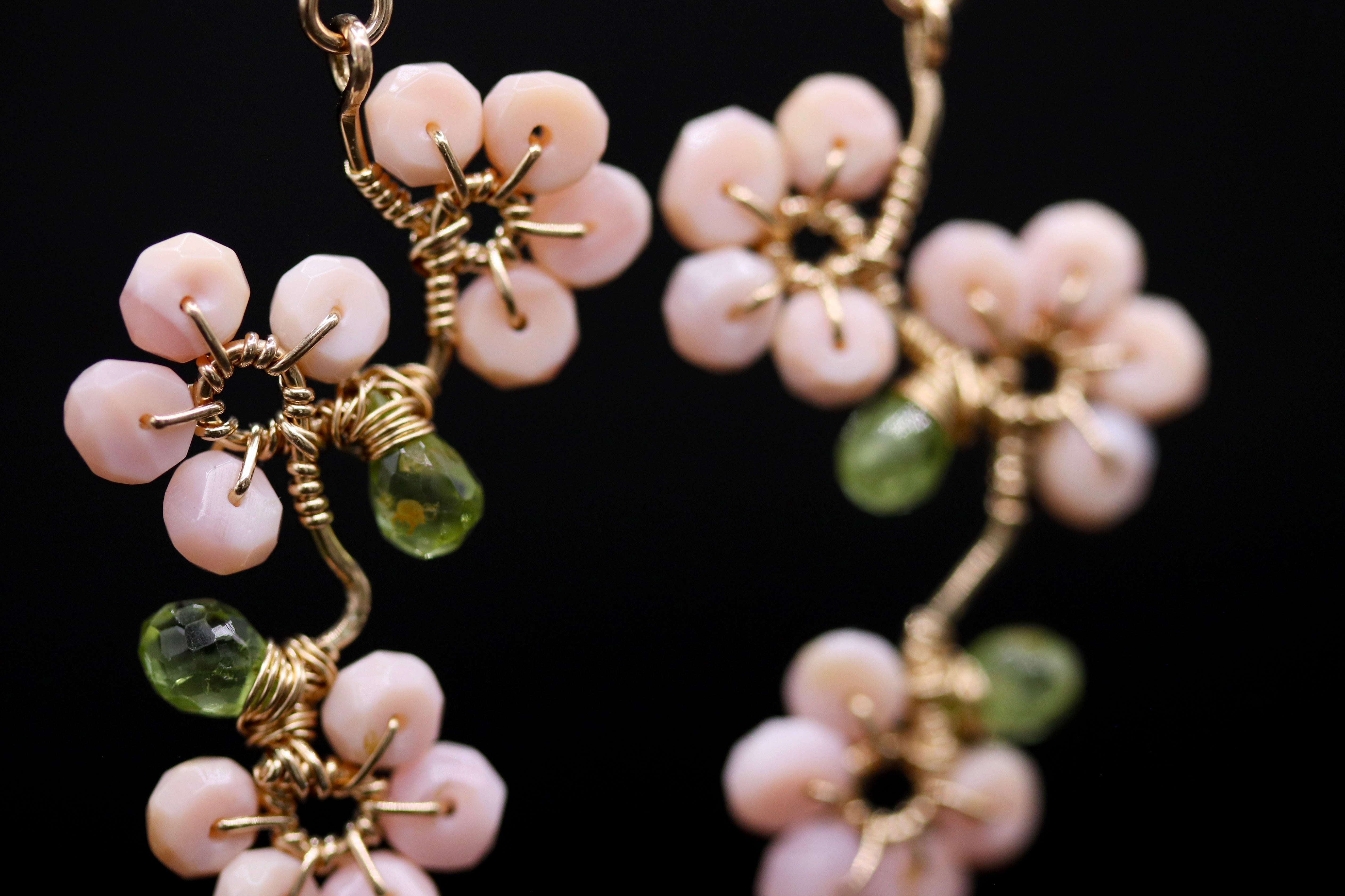 A close-up view of a pair of delicate earrings with pink and green gemstones, featuring intricate wirework and dangling from a gold-colored metal frame.
