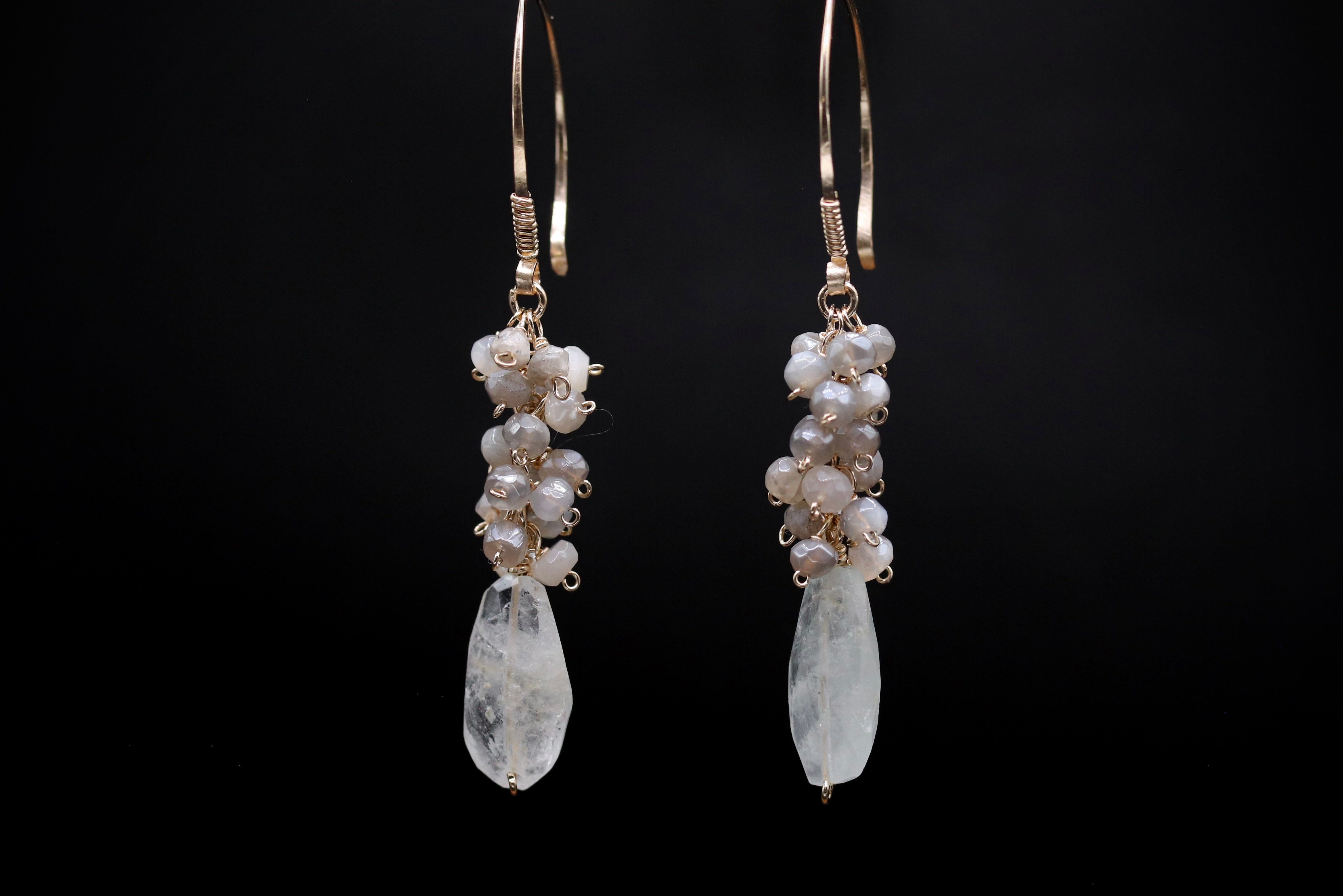 A pair of elegant earrings with clear gemstone pendants and metal hooks, set against a black background.