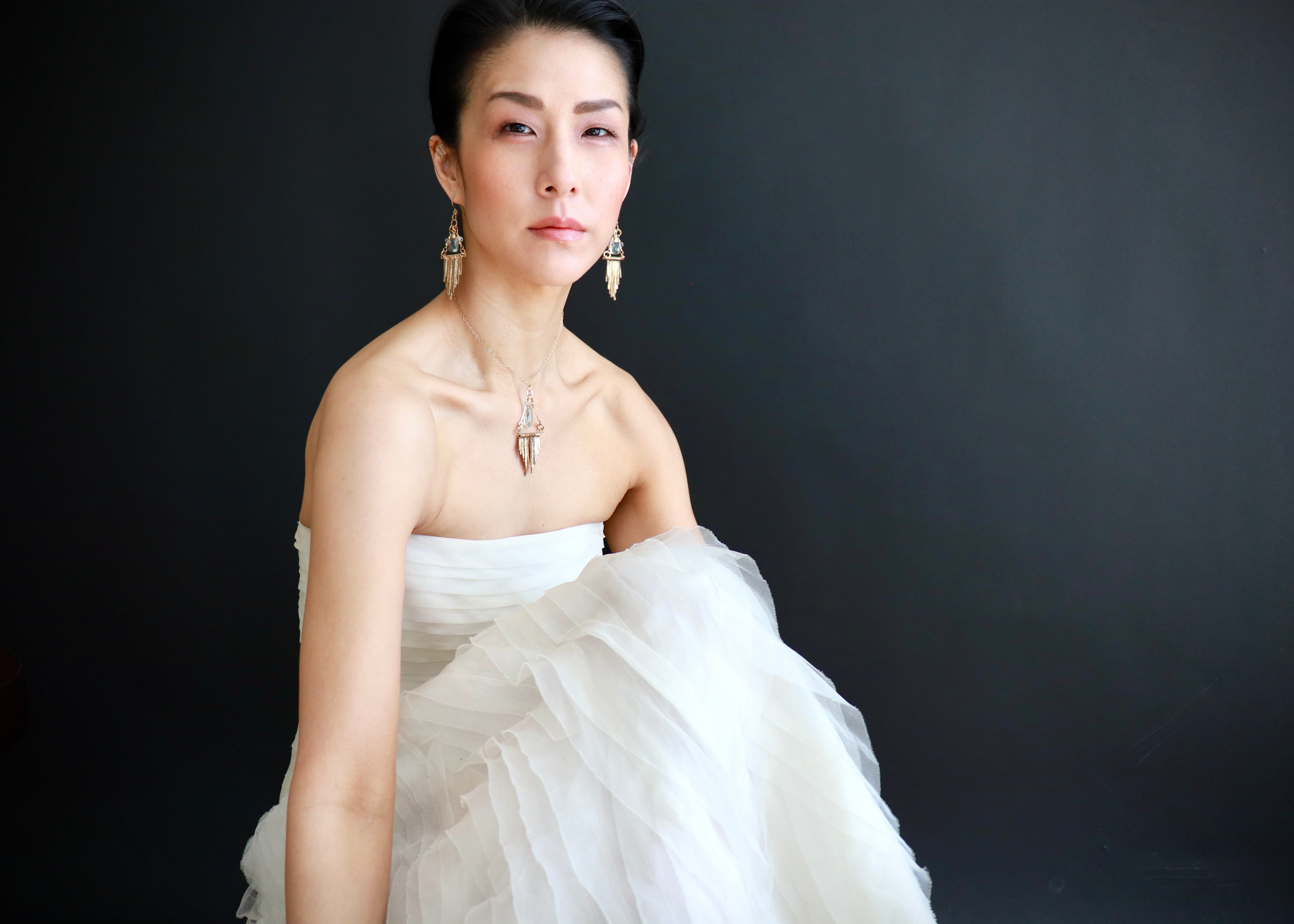 A woman in a white strapless dress poses against a dark background, wearing large earrings and looking directly at the camera.