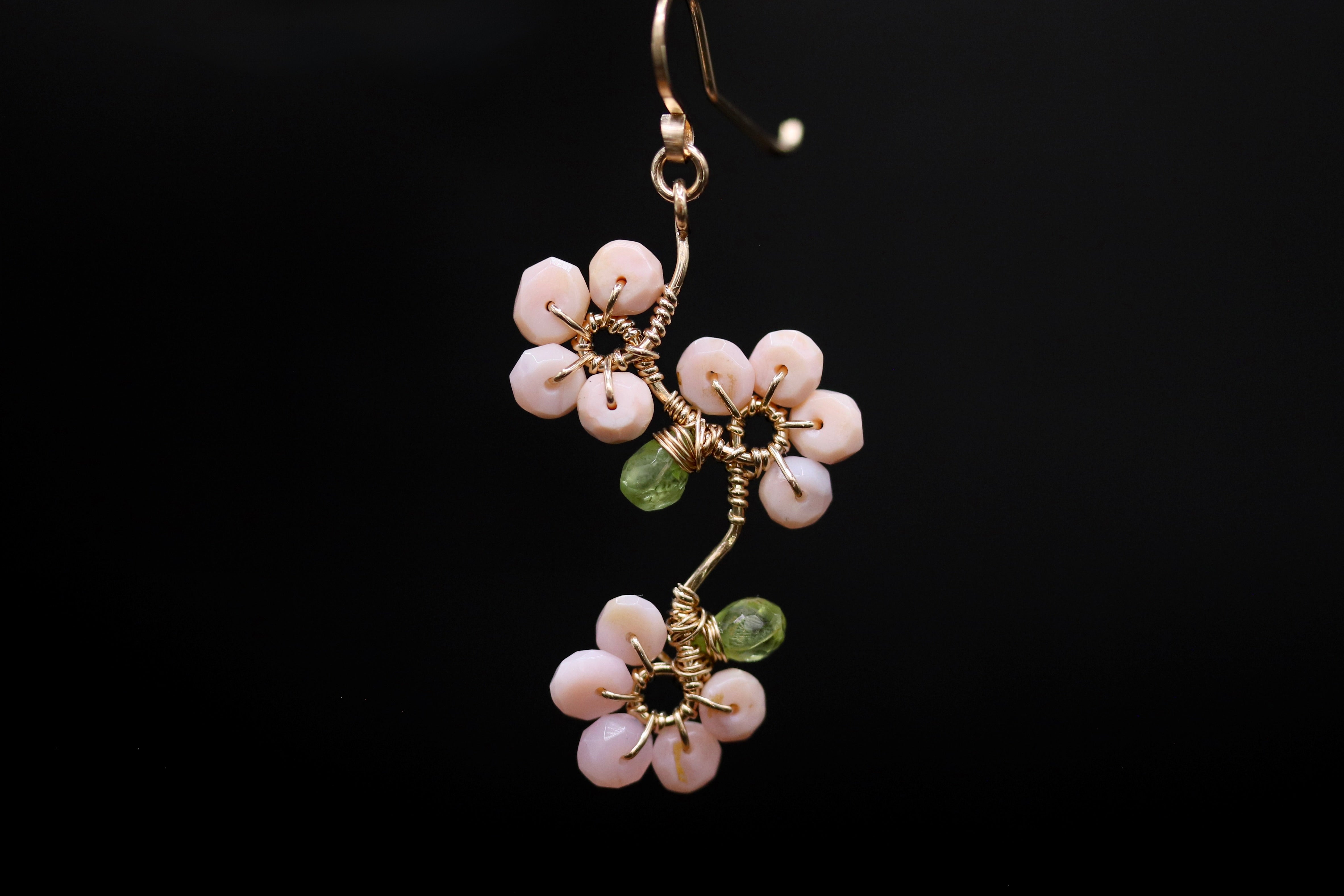 A pair of earrings with delicate pink flowers and green stones, suspended against a black background.