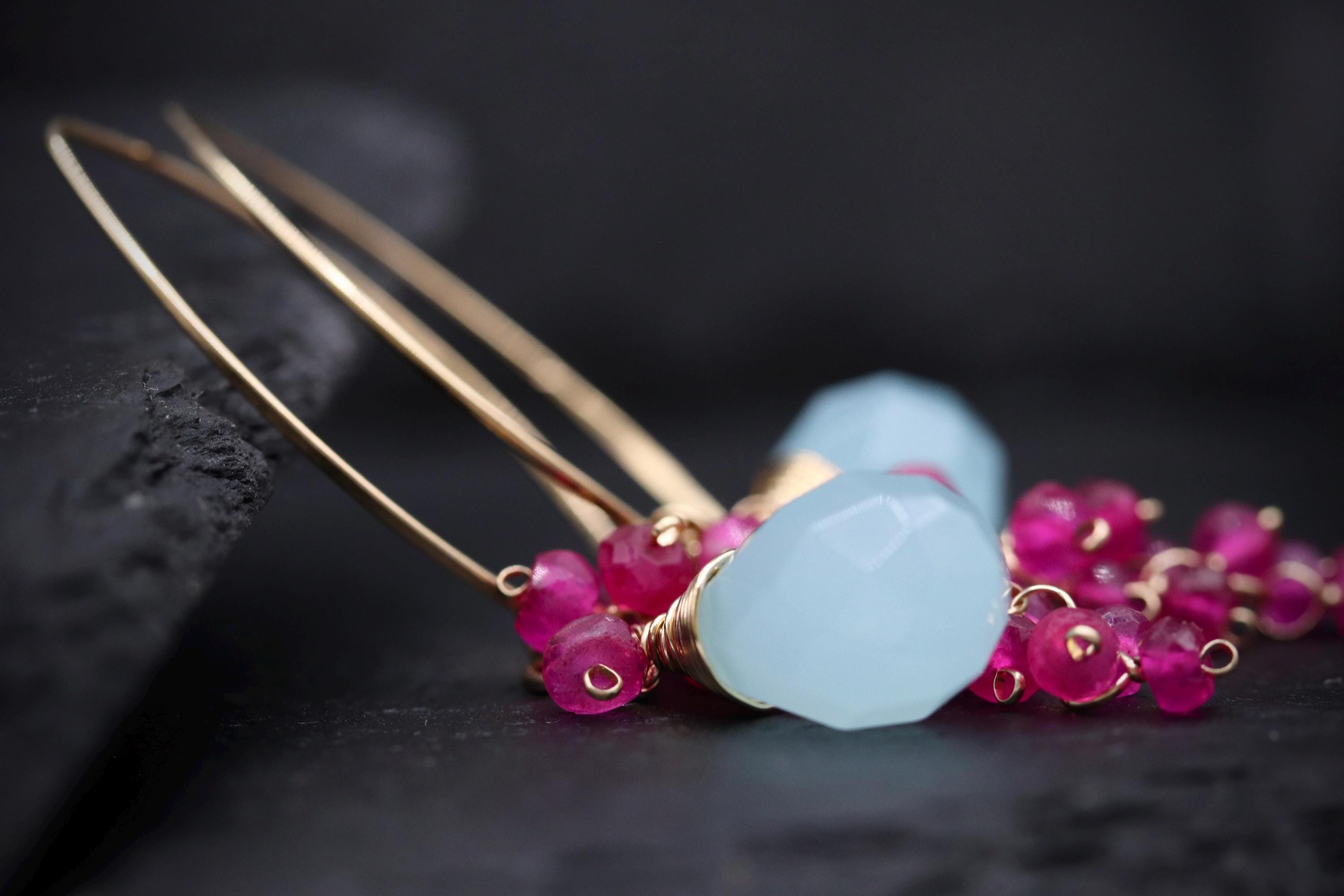 A close-up image of a pair of gold hoop earrings with blue gemstones and pink gemstones, resting on a dark surface.