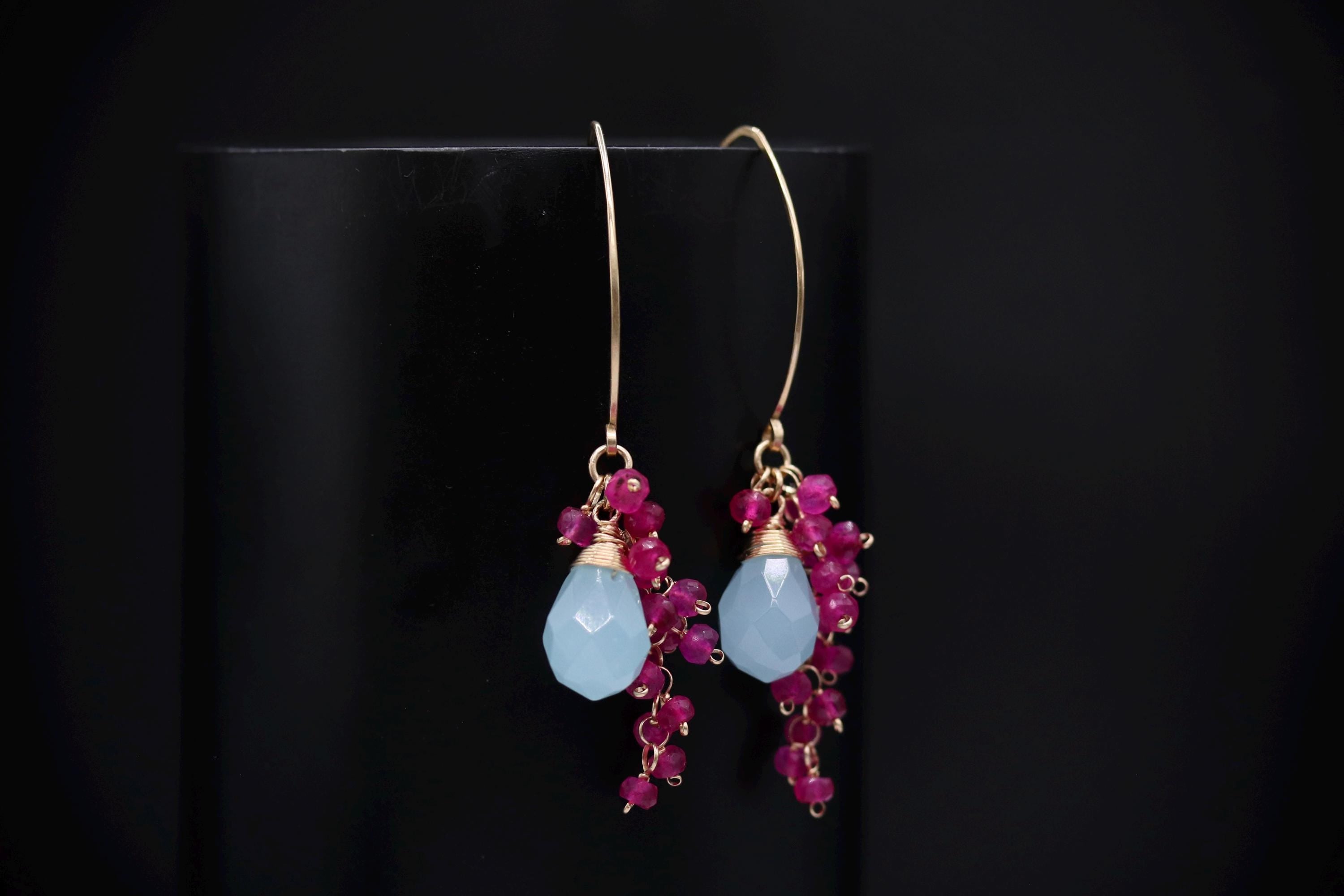 A pair of earrings with blue and pink gemstones and dangling charms, displayed against a black background.