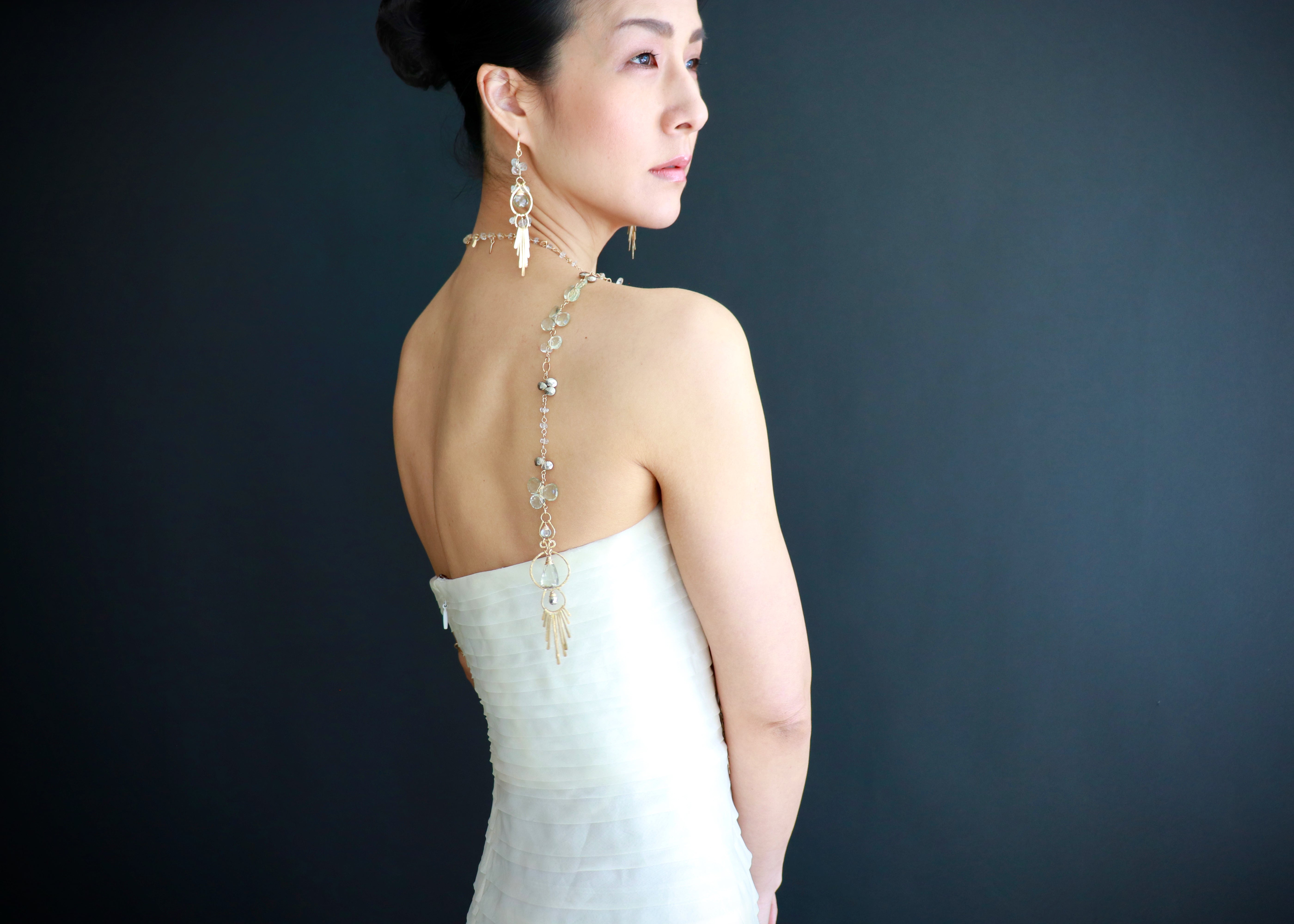 A woman in a white strapless dress with a long necklace stands against a dark background, her back to the camera.