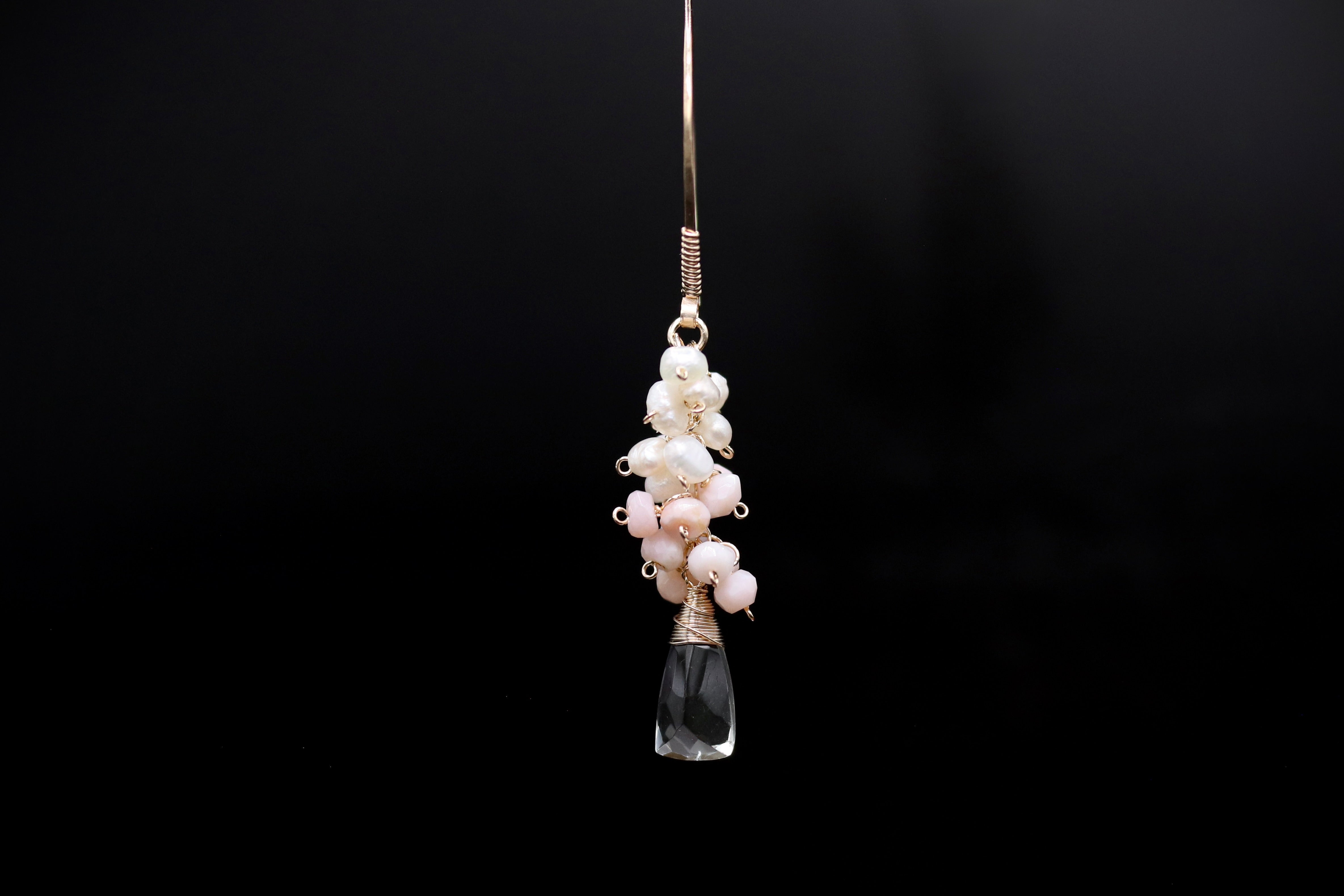 A clear glass pendant with a cluster of white and pink pearls hanging from a gold-colored metal hook against a black background.