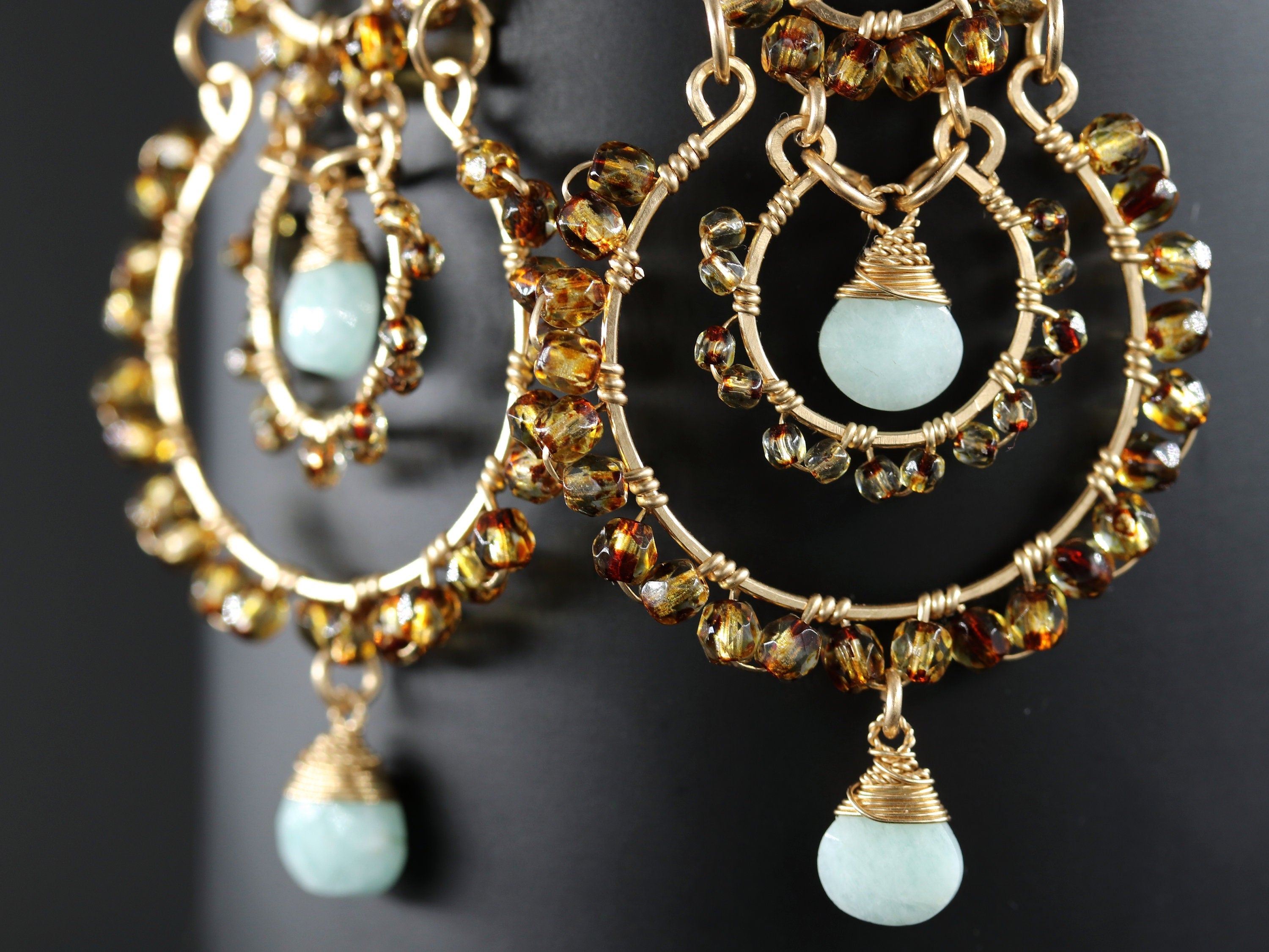 May include: Close-up of gold-toned hoop earrings with intricate wirework. The earrings feature small amber-colored beads and teardrop-shaped, pale blue-green gemstones. The design is elegant and detailed, suitable for special occasions.