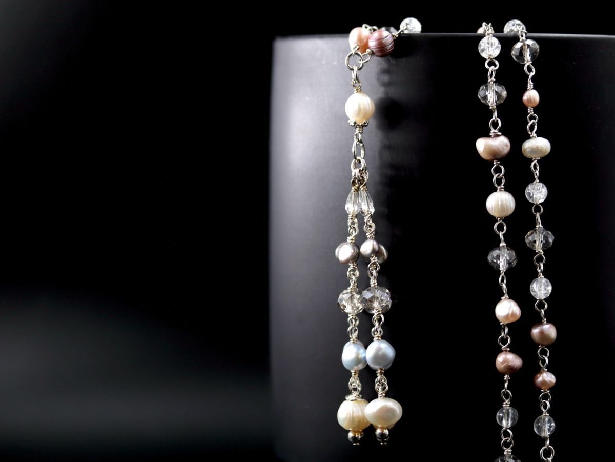 May include: Two delicate necklaces with silver chains and pearls in shades of white, pink, and blue. The necklaces are draped over a black surface.