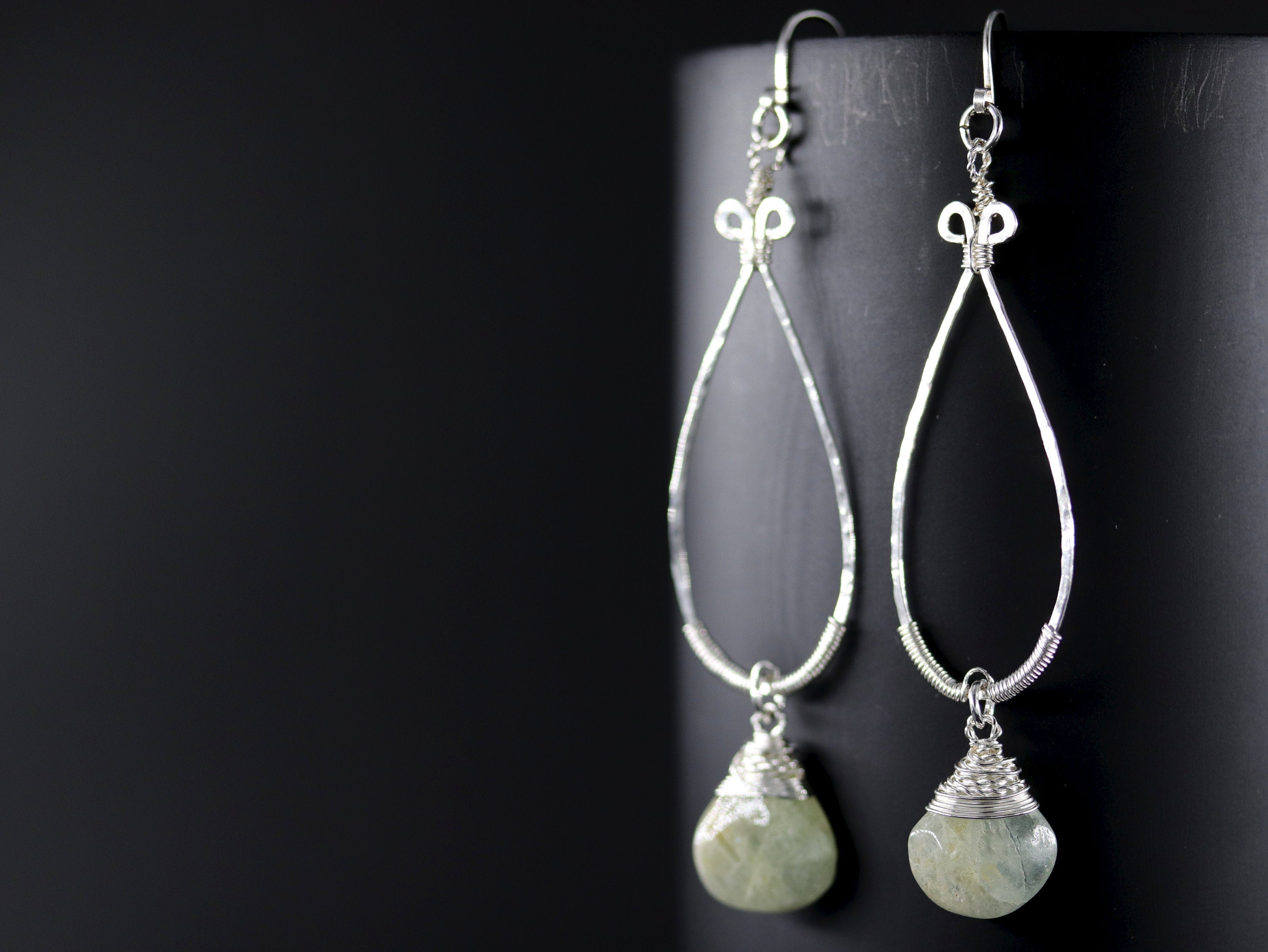 May include: A pair of silver-toned teardrop earrings with a wire-wrapped, pale green gemstone at the bottom. The earrings have a hammered texture and a decorative loop at the top, hanging against a black background.