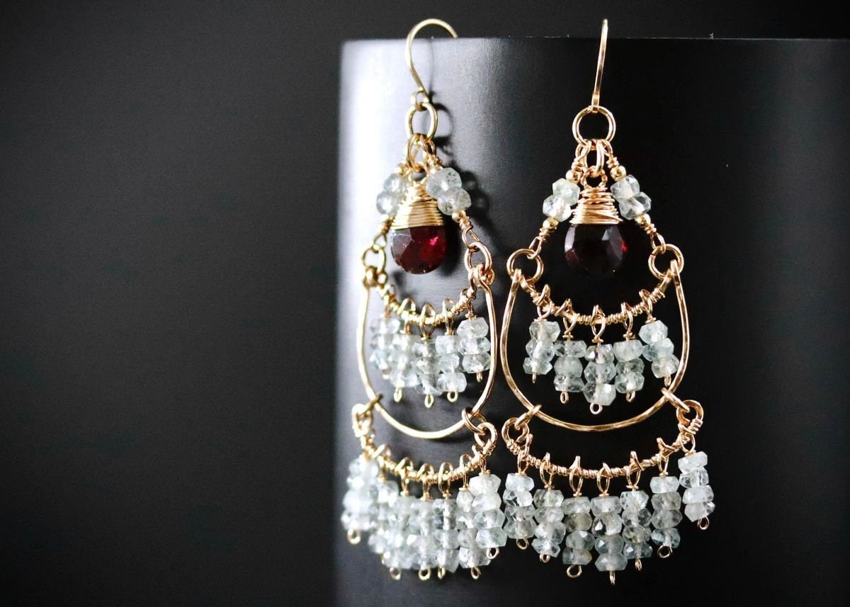 May include: Gold-toned chandelier earrings with three tiers of light blue-green beads and a deep red gemstone at the top. The earrings have a delicate, handcrafted appearance, with a dark background.