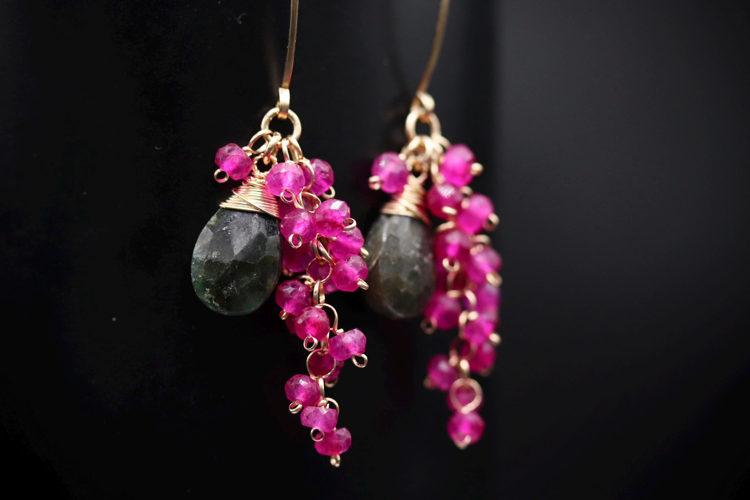 A pair of earrings with green stones and pink gemstones hanging from the ear hooks against a black background.
