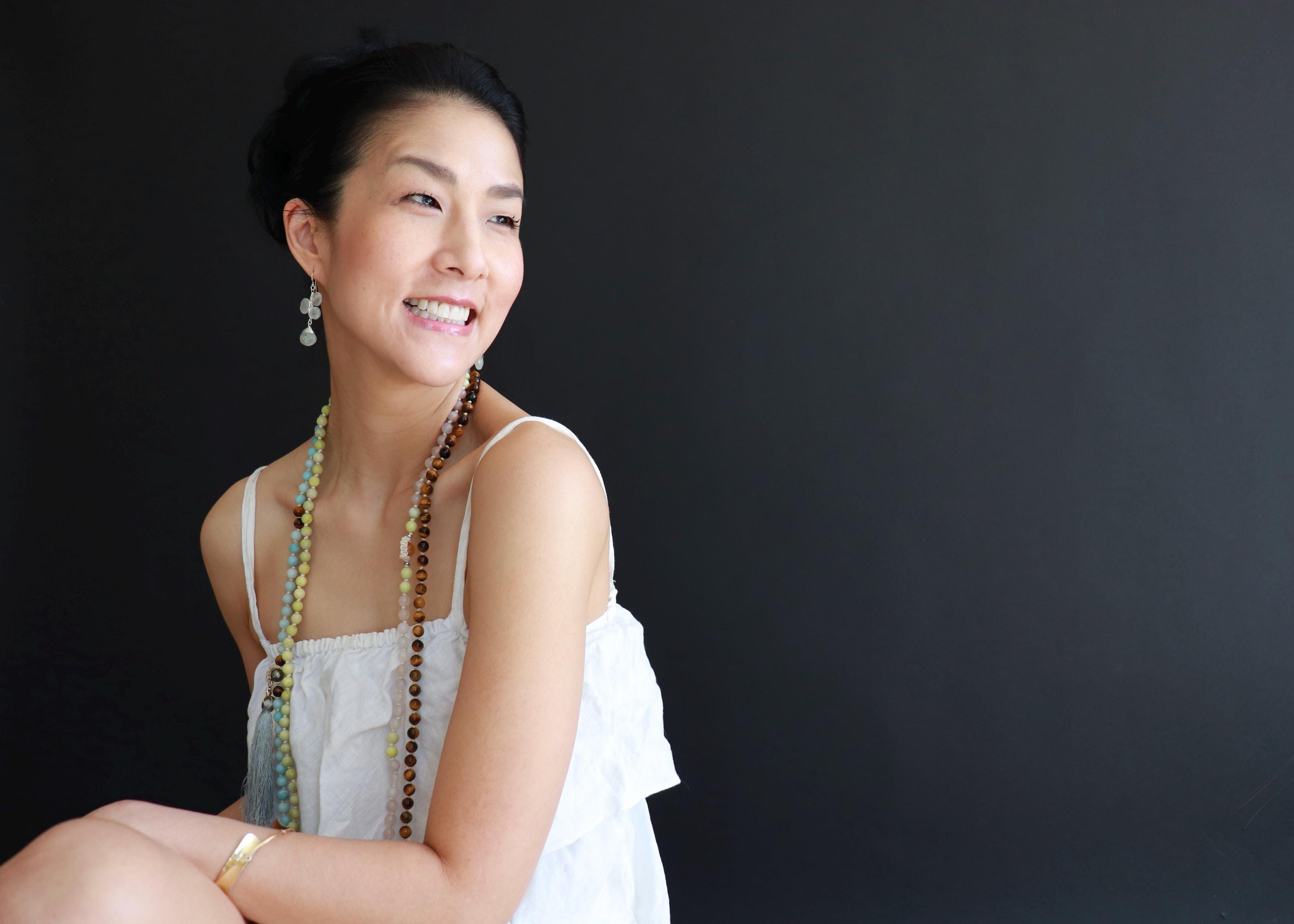 A smiling woman wearing a white top and colorful beaded necklace poses against a black background.