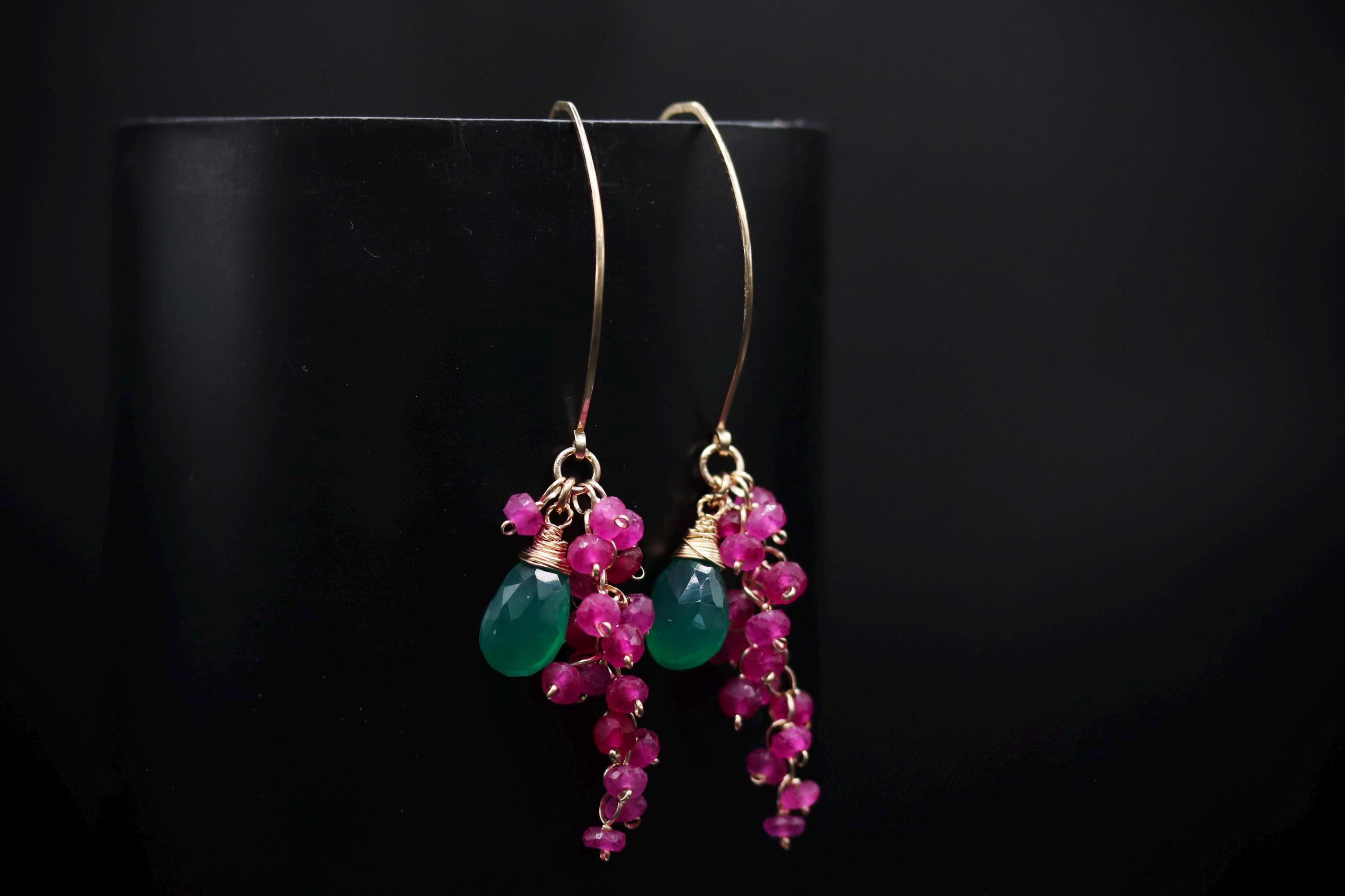 A pair of earrings with green and pink gemstones hanging from a gold hook against a black background.