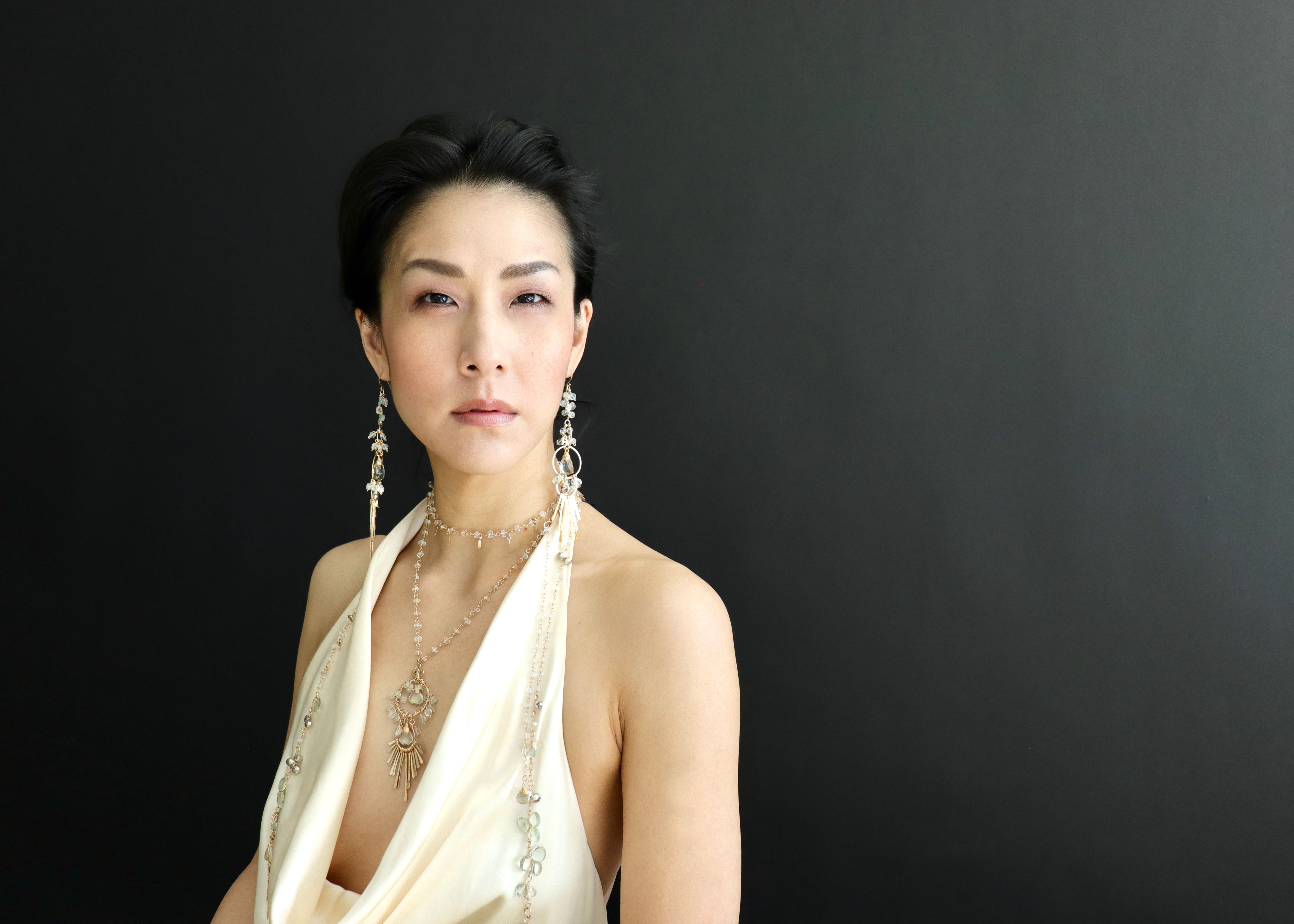 A woman wearing a white dress and large earrings poses against a black background.